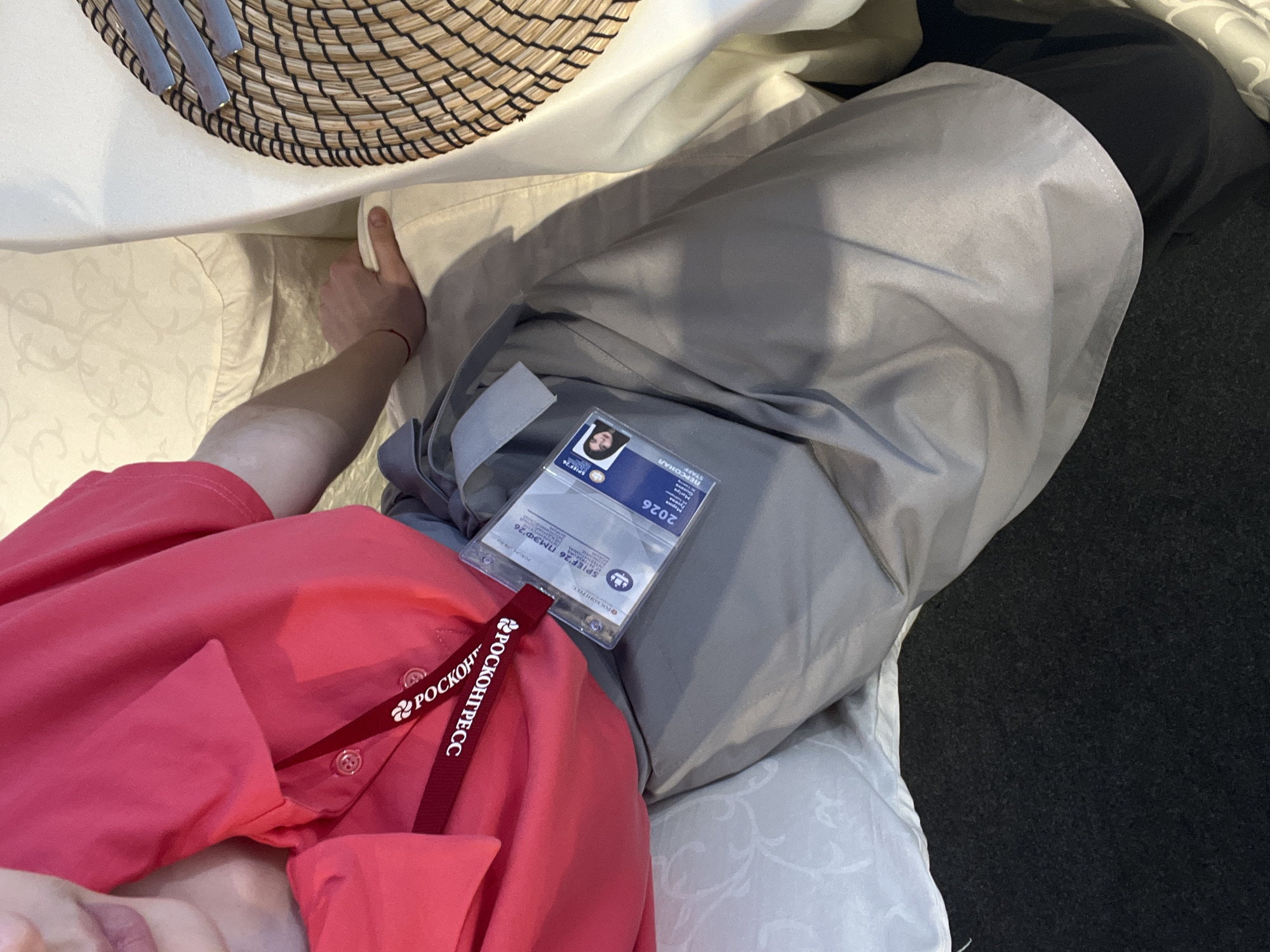 A high-angle, close-up shot captures a person seated or reclining indoors, displaying their upper torso, arm, and legs. The person wears a vibrant red polo shirt with buttons visible near the collar. A red lanyard, featuring the repeated text "РОСКОНГРЕСС" (ROSCONGRESS) and an associated logo, hangs around their neck, holding a transparent ID badge holder.
The ID badge prominently displays a small photograph of an individual, the word "ПЕРСОНАЛ" (STAFF), and the year "2026". Crucially, it identifies the event as "SPIEF'26 ПМЭФ'26", indicating the St. Petersburg International Economic Forum scheduled for 2026. The badge also includes the ROSCONGRESS logo and what appears to be a barcode or QR code.
The person's arm rests on a light-colored, ornate patterned cushion, which abuts a white surface, possibly a table, where a woven placemat or basket with cutlery is partially visible. Their legs are covered in gray fabric, likely part of an outer garment or trousers, extending towards a dark gray, textured floor or carpet. The overall setting suggests a casual yet professional environment, consistent with a participant or staff member resting at an indoor conference or event.