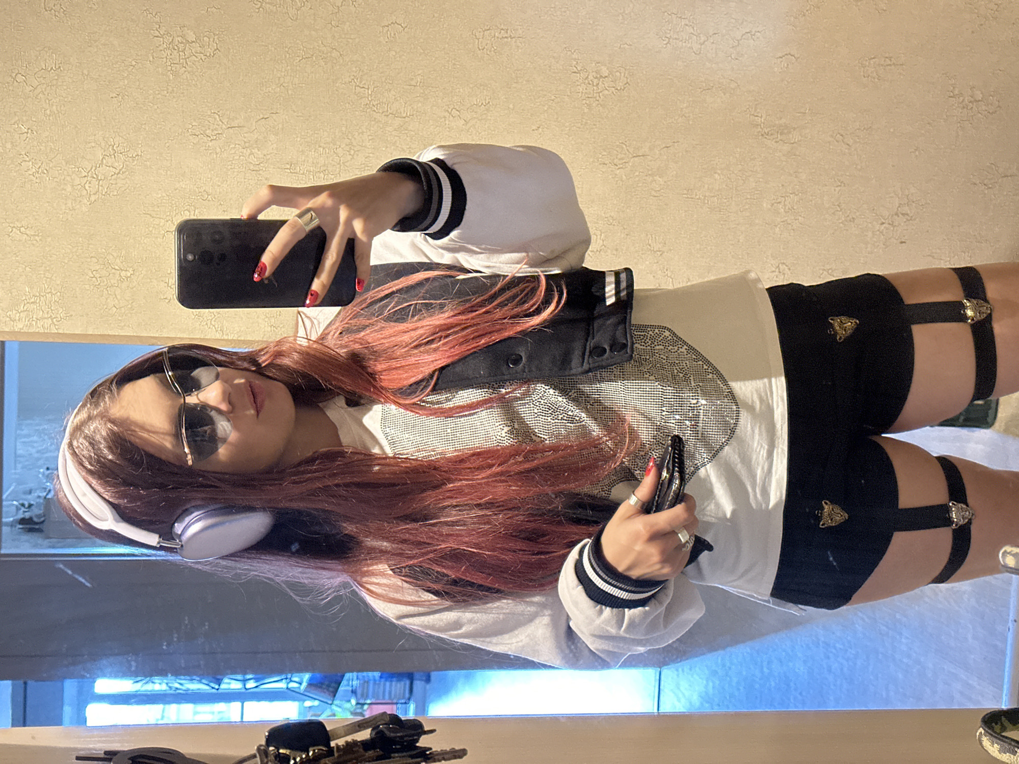 A person with long, reddish-brown hair, wearing sunglasses, a white hoodie with black trim, and black shorts with garter-like straps, takes a selfie in what appears to be a bathroom or dressing area. The person is holding a black smartphone in their right hand and a black object, possibly a stylus or pen, in their left hand. They are also wearing white headphones around their neck. The background features a textured, light-colored wall and a reflective surface, likely a mirror, which shows the person and their surroundings. The lighting suggests it might be daytime, possibly indoors with artificial light. The overall mood seems casual and self-aware. There is no visible text in the image.