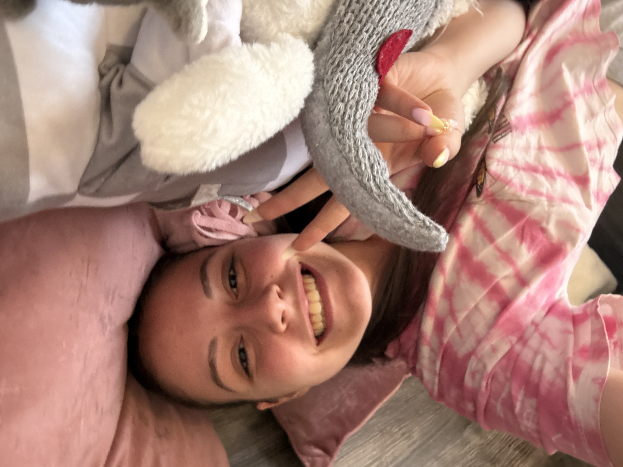 A young woman is lying down on her back, smiling widely and looking up at the camera, surrounded by plush toys and soft furnishings. She is making a peace sign with her right hand, positioned near her face, and her left hand is also visible, holding what appears to be a small, beaded ring. Her cheerful expression is characterized by a broad smile, revealing her teeth, and bright eyes.
She is wearing a pink and white tie-dye top. Her fingernails are painted with a light pink base and yellow/gold tips. To her left, a large, fluffy white plush toy is visible, and to her right, a gray knitted plush toy with a small red felt accent. Her head rests on a pink pillow or blanket, and the background suggests a wooden floor or a similar indoor surface. The setting appears to be a cozy indoor environment, likely a bedroom, creating a relaxed and playful atmosphere. The image captures a moment of lightheartedness. This scene is set in Bulgarovo, Bulgaria.
