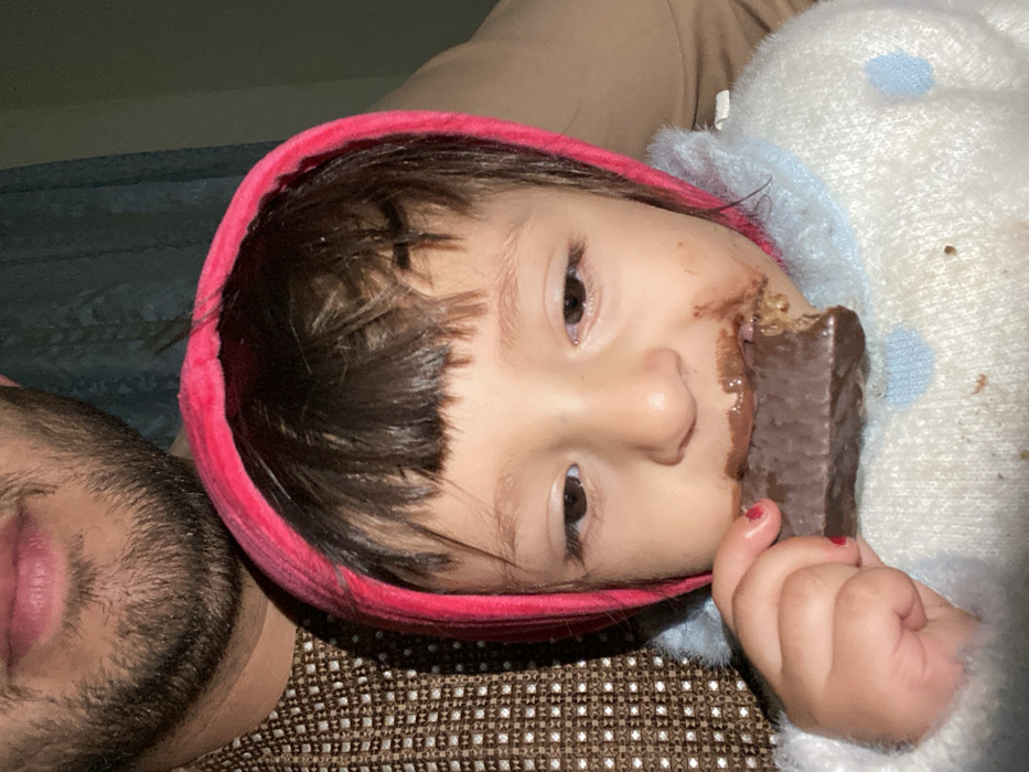 In Kandahar, Afghanistan, a close-up image depicts a young child, possibly female, actively eating a chocolate bar. The child wears a bright pink hood and a white knitted sweater with light blue polka dots, exhibiting chocolate smudges around the mouth and on the sweater. The child's left hand, with red nail polish on a fingernail, holds the partially consumed rectangular chocolate bar. In the blurred background, a partial view of an adult male's lower face, featuring a beard, and a brown patterned garment are visible. The background also shows a blue curtain and a plain light brown surface, indicating an indoor domestic setting.