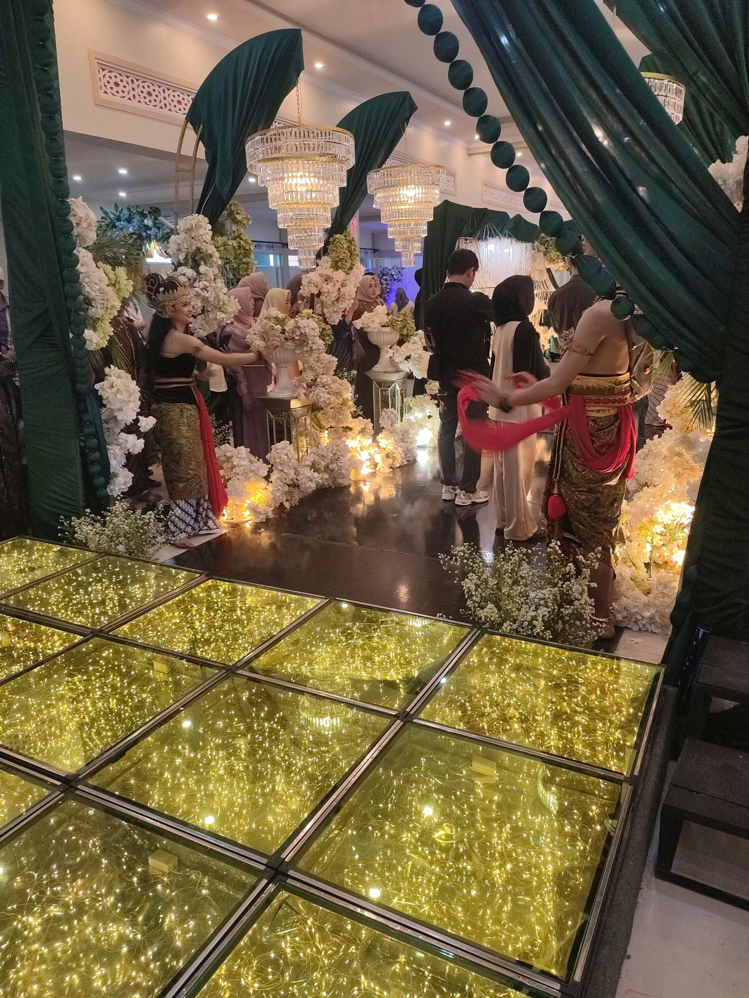 The image depicts a festive indoor scene, likely a wedding reception, located in Karangmalang, Indonesia. A glass-covered dance floor, illuminated by numerous warm-toned string lights, occupies the foreground. In the mid-ground, individuals dressed in traditional attire, featuring ornate headpieces and colorful fabrics, are engaged in what appears to be a cultural performance or a ceremonial procession. One performer, adorned in a dark top and a patterned skirt with a red sash, is gracefully holding a bouquet of white flowers. Another performer, wearing a golden skirt and a red sash, is also visible. Several other guests, some in veiled attire, are observing the performance. The background is decorated with lush green drapery, large crystal chandeliers hanging from the ceiling, and abundant white floral arrangements, creating a luxurious and celebratory atmosphere. The overall scene suggests a cultural event taking place in a well-decorated venue.