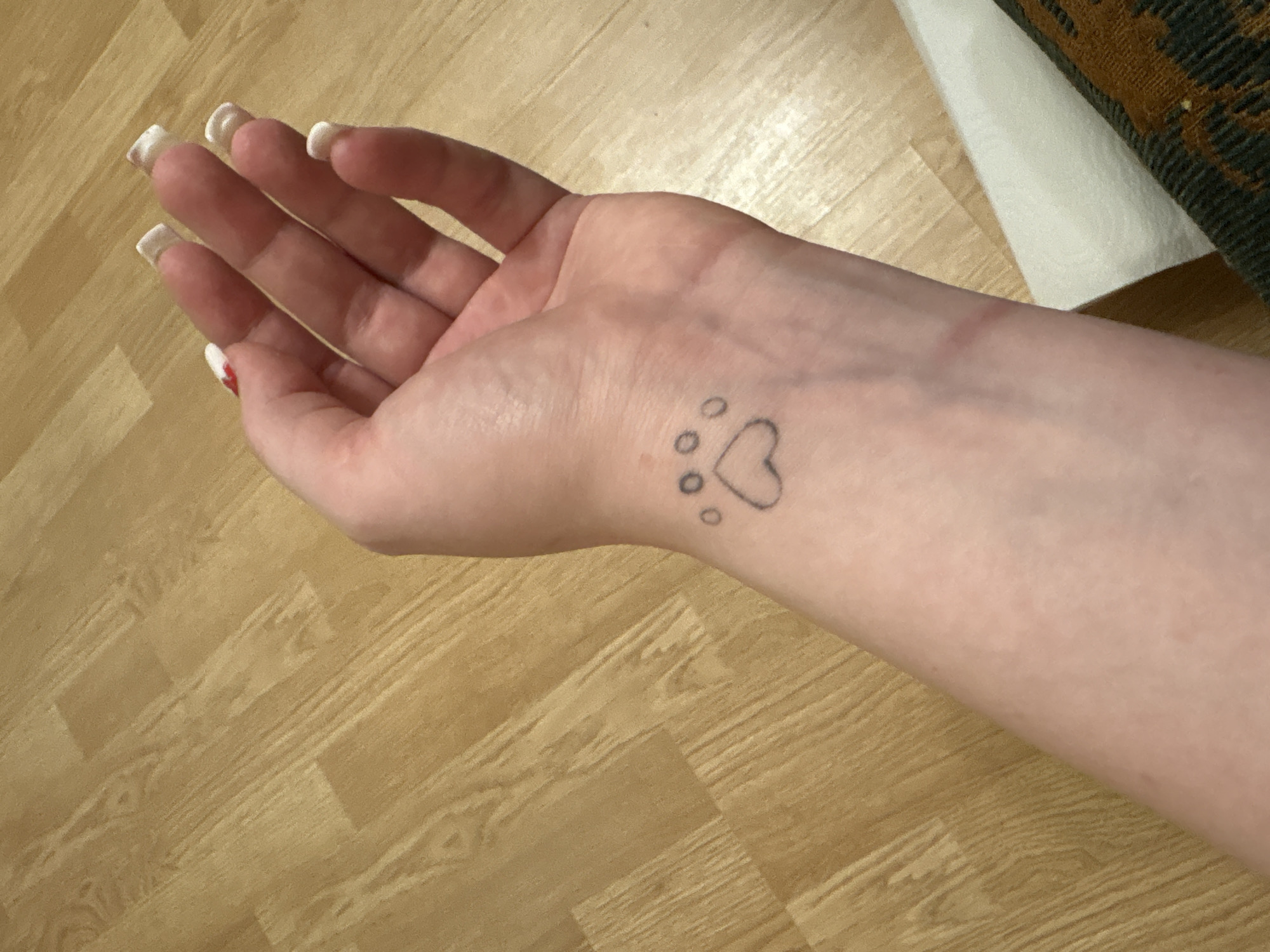 A Caucasian hand and wrist are shown against a background of wood-look flooring. A tattoo of a paw print incorporating a heart is visible on the wrist. The fingernails on the hand are painted with a white French manicure and one finger has a red design on it. The lighting is bright and diffused. A white piece of fabric and a dark-patterned object are visible near the arm. The location is not identifiable as Izhevsk, Russia based on the visible elements.