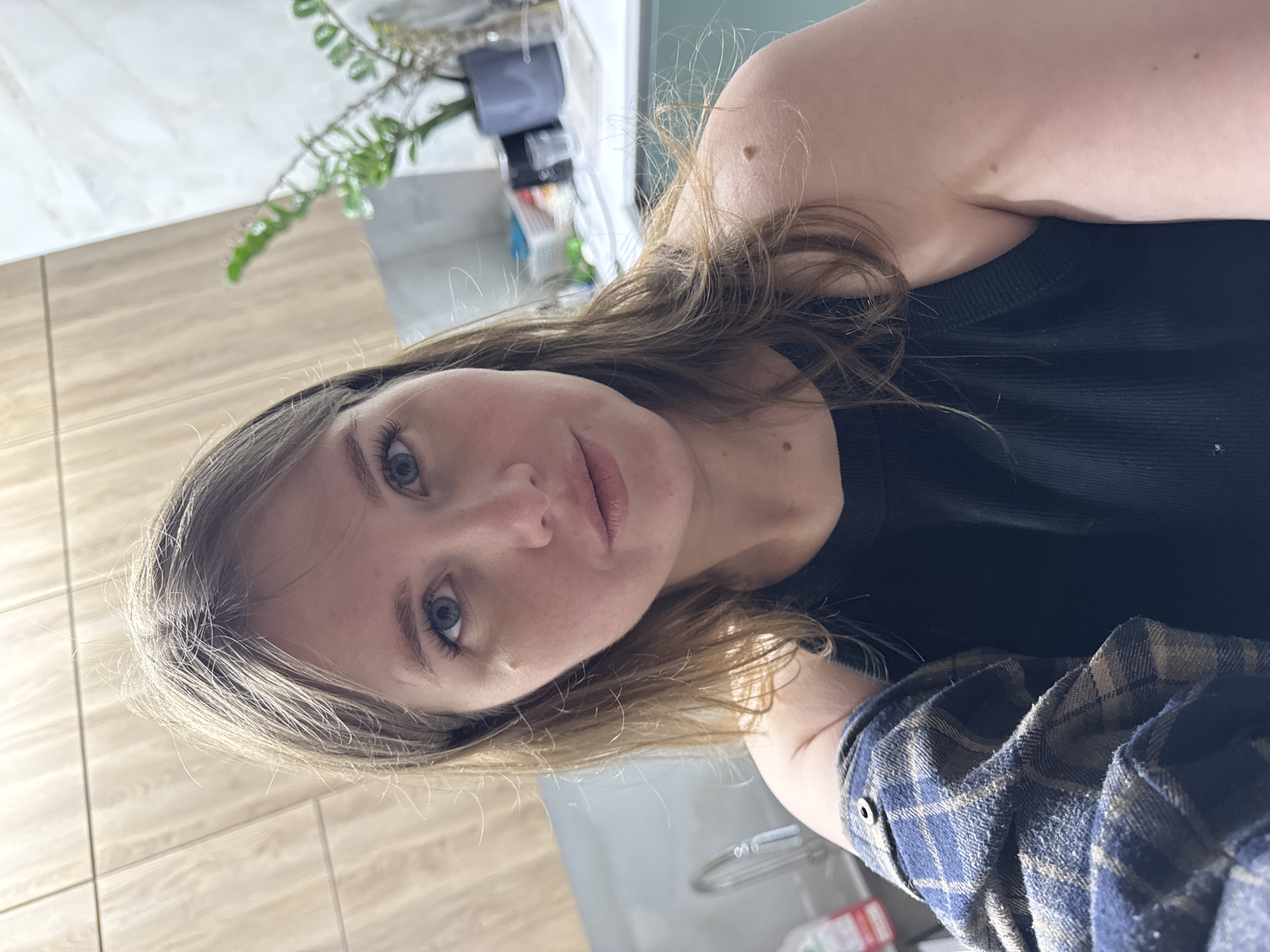 A young woman with light hair and blue eyes is posing for a selfie. She is wearing a black tank top and a blue and brown flannel shirt. The background appears to be a kitchen, with wooden cabinets, a plant, and other kitchen items visible. The location is specified as Gornyi (Dvurechenskoe S/P), Russia.