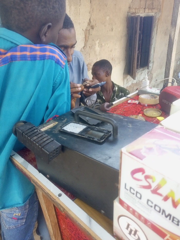 An outdoor mobile phone repair or sales operation is depicted in Akwanga, Nigeria. Three males are gathered around a table covered with a red patterned cloth. The male in the center, wearing a grey shirt, is focused on a small electronic component held in his left hand while also holding a smartphone in his right hand. To his right, a younger male in a green patterned shirt holds an open flip phone. Partially visible on the left, a third male in a light blue shirt and jeans observes the activity. On the table, various tools and items are present, including a dark portable electronic device with a handle, disassembled phone parts (screens or keypads), a white power strip, adhesive tape, small containers with unknown contents, and a red object marked "LEER." In the immediate foreground on the right, a stack of cardboard boxes is visible, with the top box displaying "CSLNM" and "LCD COMBO." The background features a textured, light-colored wall with a dark, barred window.