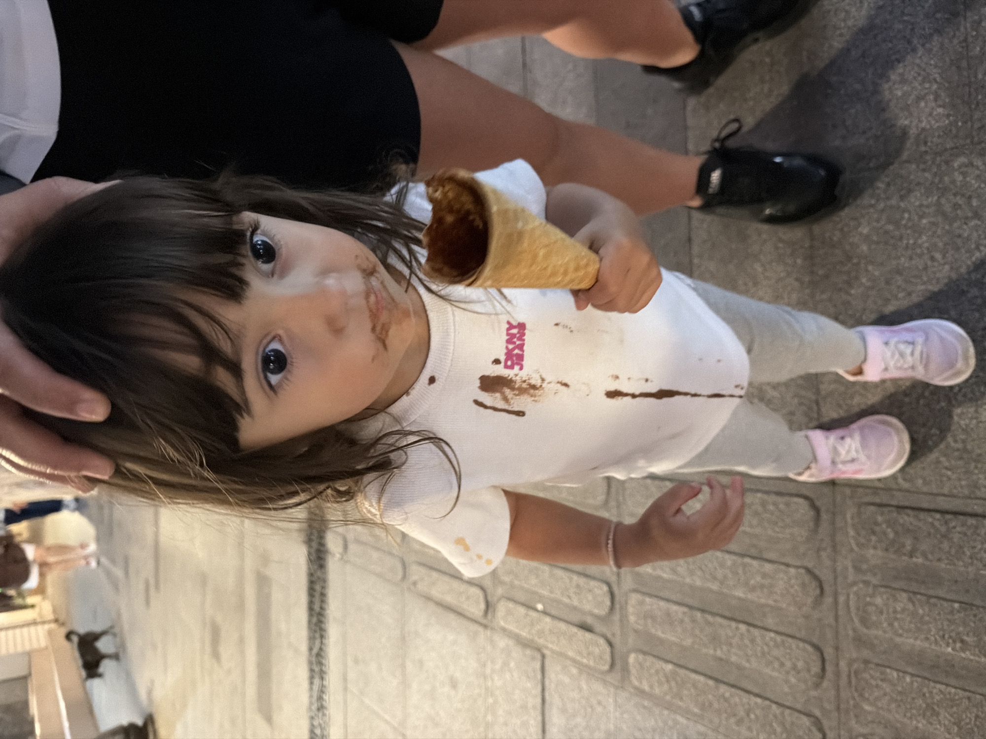 A young child with dark hair and big eyes is standing on a cobbled street. The child is wearing a white t-shirt with chocolate stains and pink sneakers. The child is holding a partially eaten chocolate ice cream cone. A person's hand is resting on the child's head. In the background, there are blurred figures of people and a small dog. The scene appears to be outdoors in an urban environment, possibly Burgas, Bulgaria, given the cobblestone pavement which is typical of the city's central areas and the Sea Garden.