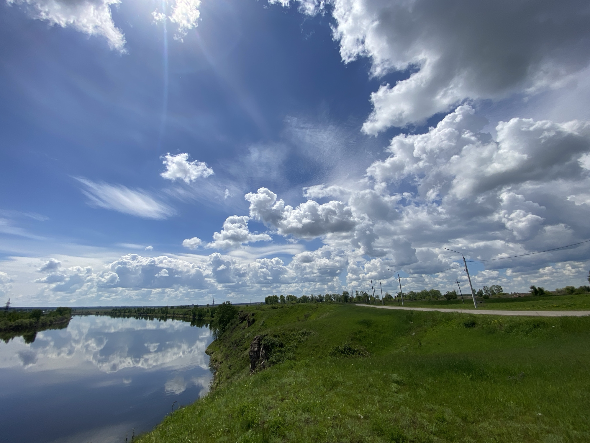 This media file presents a wide, sunlit landscape in Novomaltinsk, Russia, characterized by a dynamic sky, a calm river, and lush green terrain.
The sky occupies a significant portion of the image, displaying a vivid blue canvas adorned with various cloud formations. In the upper left, the sun casts a bright lens flare, indicating a clear, bright day, likely midday. Wispy, thin cirrus clouds streak across this part of the sky, while the right side is dominated by large, fluffy cumulus clouds, some appearing brighter white and others with darker, grayer undersides, suggesting depth and thickness. The clouds are clearly reflected on the smooth surface of the river.
The wide river stretches across the middle ground, its dark, reflective water acting as a mirror to the expansive sky above. The reflections of the blue sky and white clouds are remarkably clear and sharp, almost creating a symmetrical composition with the actual sky. On the far bank of the river, a line of trees is visible, extending into the distance.
In the foreground and to the right, a verdant grassy bank rises steeply from the river's edge, transitioning into a gently sloping terrain. The grass is a vibrant green, suggesting healthy growth. A paved road runs along the top of this bank, curving slightly into the distance on the right side of the frame. Alongside the road, several utility poles with power lines are visible, providing a hint of human infrastructure within the natural setting.
There are no people or visible text in the image. The scene conveys a sense of peaceful serenity and the vastness of nature under a bright, cloud-dotted sky.