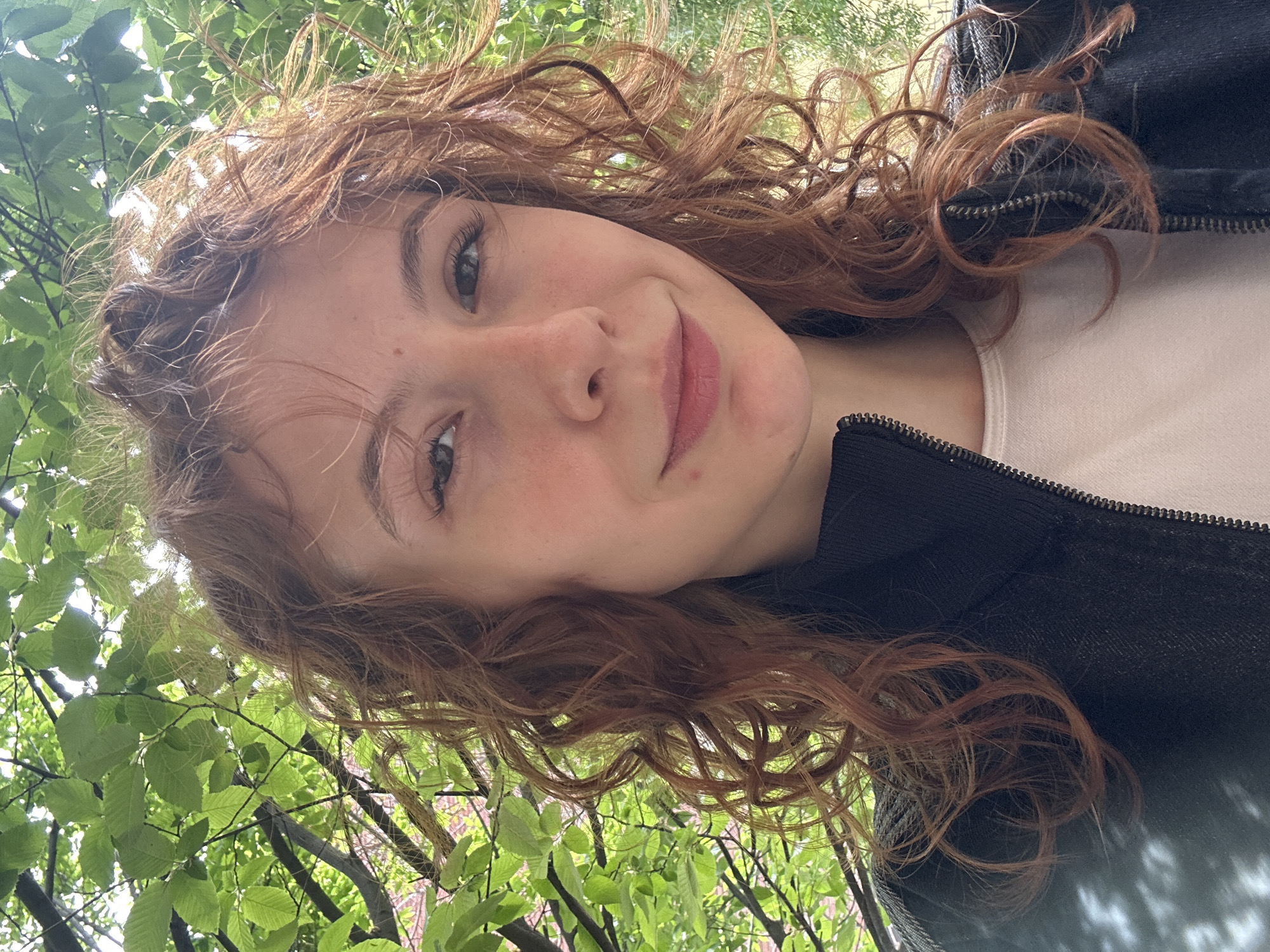 A selfie captures a young person, likely female, outdoors under a canopy of green trees in what appears to be Novi Sad, Serbia. The person looks slightly upwards towards the camera with a gentle, faint smile. Their light-colored eyes, possibly green or hazel, are visible. They have abundant reddish-brown, curly hair that frames their face and extends around their shoulders.
The individual is wearing a black jacket with a visible zipper, layered over a light-colored top. The background is dominated by vibrant green leaves, suggesting a sunny daytime setting, with natural light filtering through the foliage. The scene conveys a relaxed and pleasant outdoor moment. No text is visible in the image.