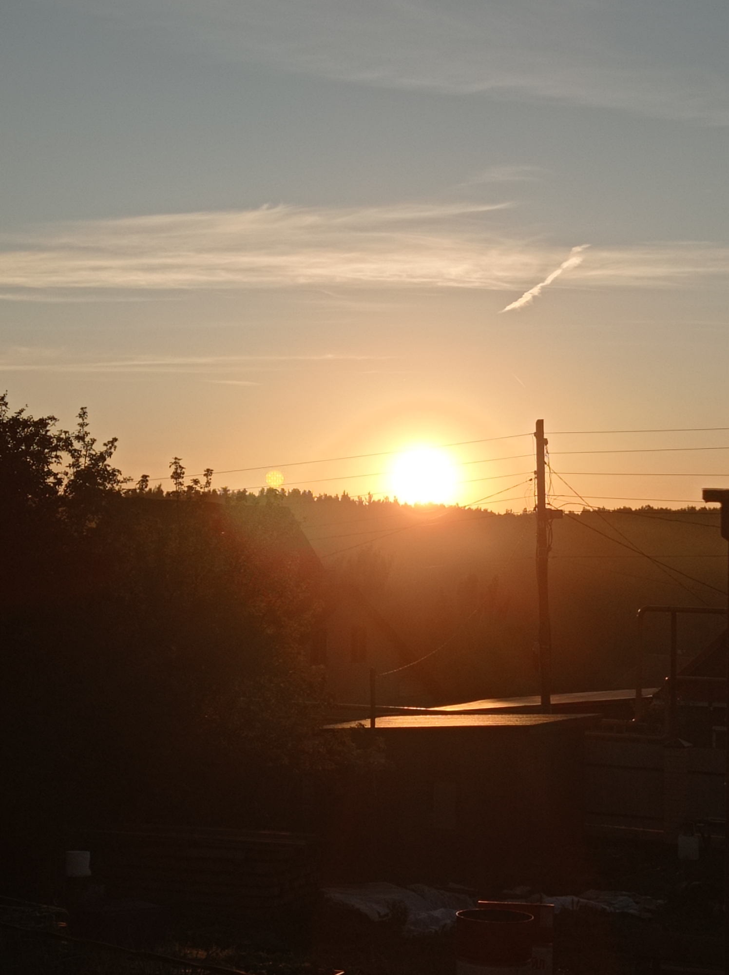 A bright, setting sun casts a warm glow over a rural landscape. The sun is positioned in the center of the frame, with rays of light extending outwards. A silhouetted line of trees forms the horizon. In the foreground, a power pole with several wires is visible on the right, and the dark silhouettes of trees and some structures are present on the left and bottom of the image. The sky is a gradient of pale blue to orange. The scene appears to be captured in Degtyarsk, Russia.