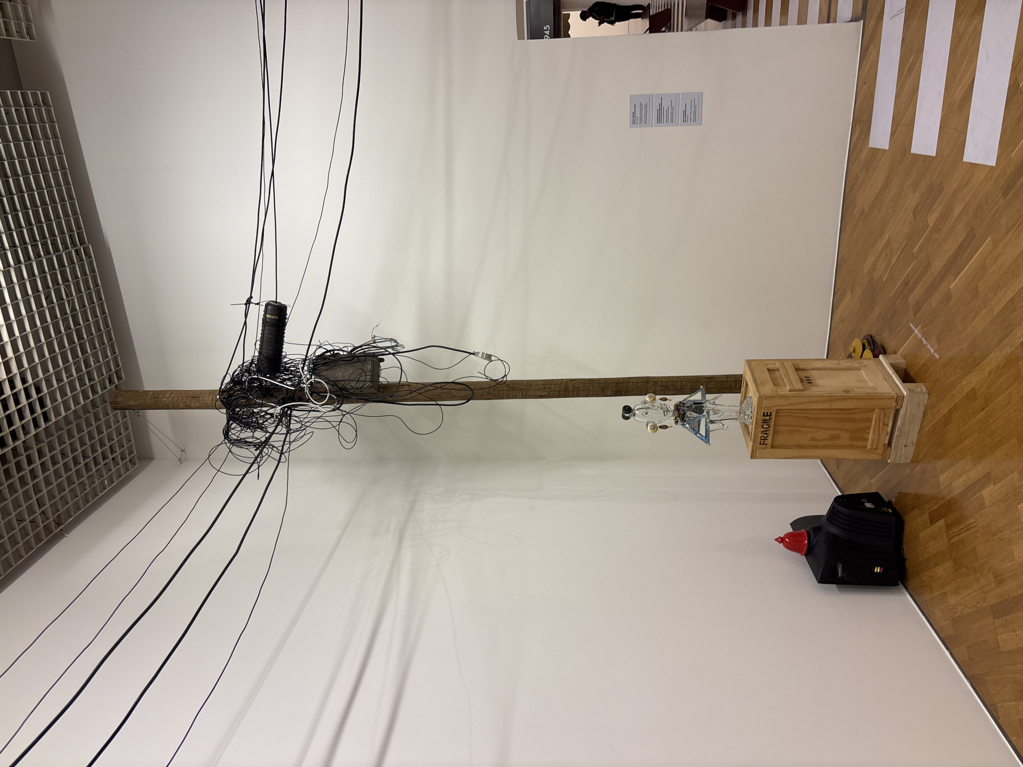 This indoor shot shows an art installation against a white wall and a parquet wooden floor. The installation features a wooden beam fixed horizontally, with a chaotic nest of black wires and equipment at one end and a wooden crate containing a clear glass and metal structure at the other. A black bag with a red cone-shaped object sits on the floor near the crate.
On the upper right portion of the wall, there's artwork depicting a person, along with some texts mounted on the wall. The image suggests a gallery or museum setting. The city of São Paulo, Brazil, is not identifiable from the visual information provided.