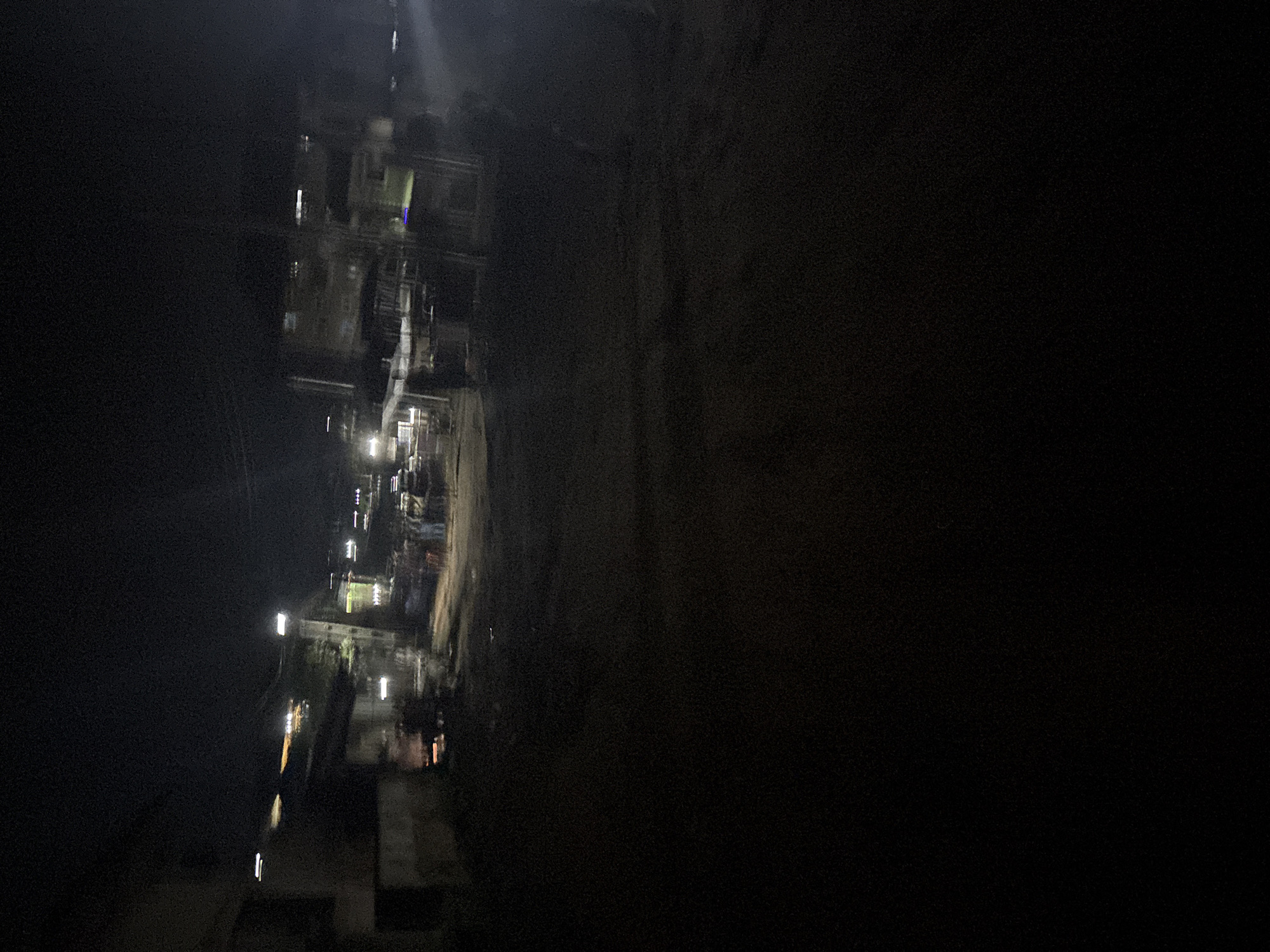 The image depicts a dimly lit street scene at night in Nkpor, Nigeria. The foreground is dark, obscuring most details. In the mid-ground, faint lights from buildings and street lamps illuminate a road or pathway. Several figures, appearing as indistinct silhouettes, are visible on the right side of the scene, gathered near what might be stalls or small structures. The background shows more buildings with illuminated windows, suggesting a residential or commercial area. The overall impression is one of a busy, albeit dimly lit, urban environment at night.