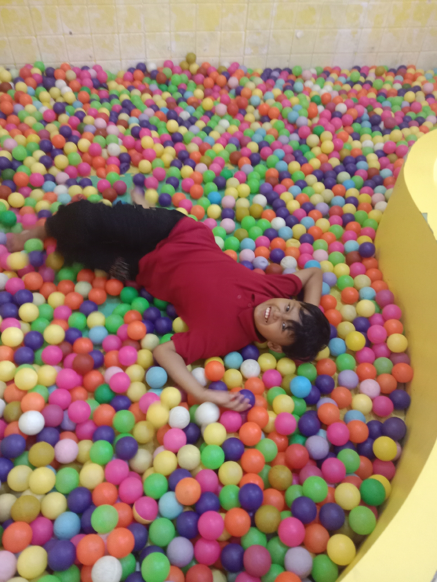 A young boy with dark hair, wearing a red shirt and black shorts, is lying on his back in a ball pit filled with brightly colored plastic balls. He is smiling at the camera and has his right arm extended upwards. The ball pit appears to be located indoors, with a yellow tiled wall in the background. The overall scene suggests a playful and recreational setting.