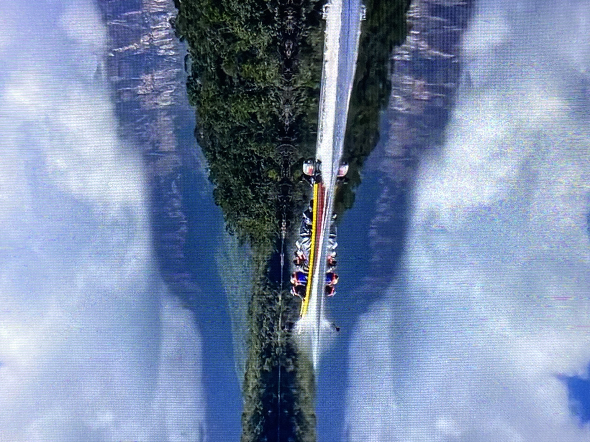 The image shows a roller coaster in Tinaquillo, Venezuela, viewed from above and reflected for symmetry. The roller coaster car, carrying multiple visible riders, travels along a white track that bisects the image vertically. To the sides of the track, there is dense green foliage, suggesting a wooded area. The background is composed of a bright, cloud-filled sky reflected below, creating a symmetrical and dream-like effect.
