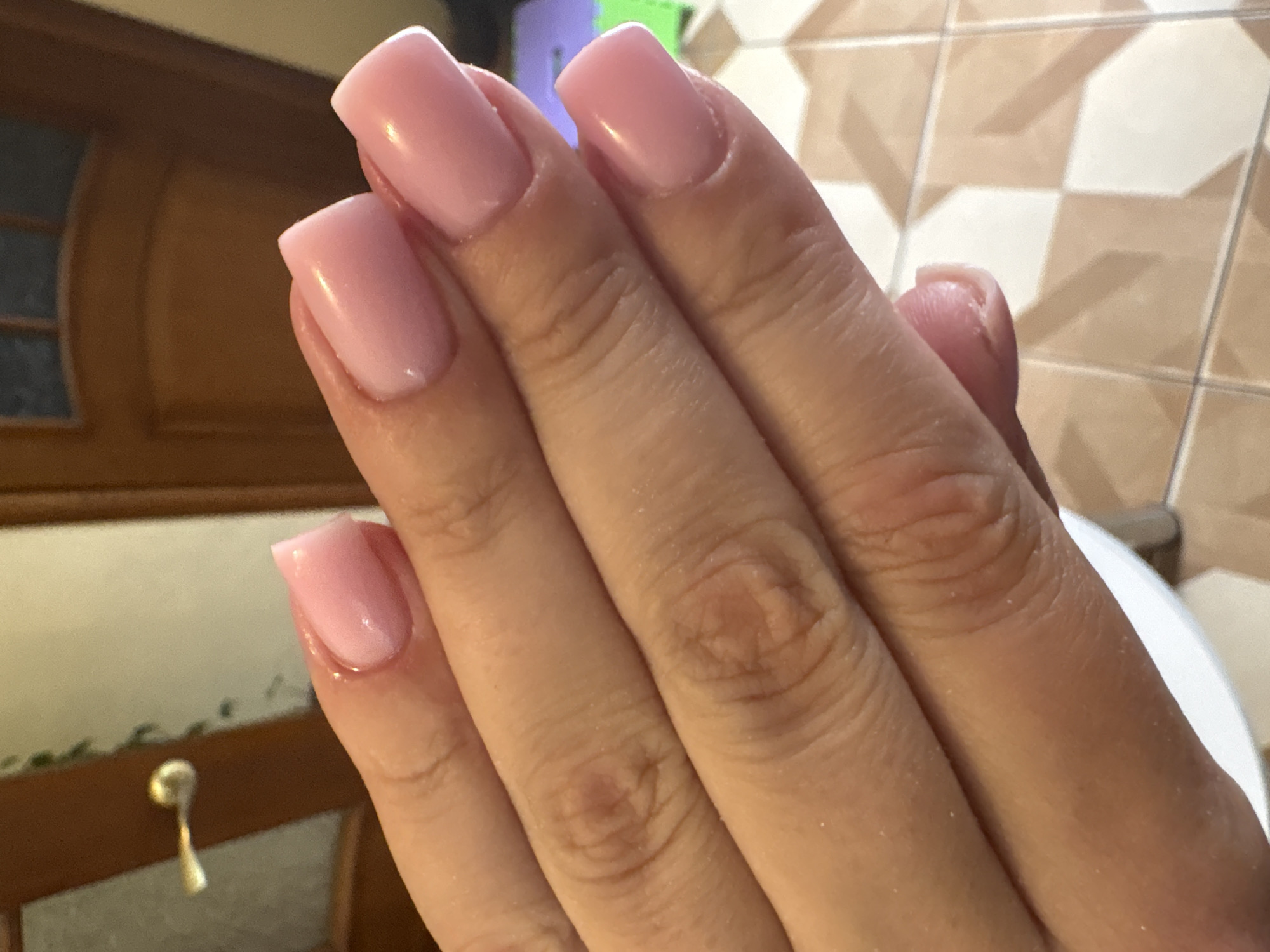 The media file displays a close-up view of a person's hand with neatly manicured, light pink nails. The nails are square-shaped with rounded edges, and appear to be acrylic or gel extensions. The skin on the hand is fair, and the fingers are shown gently curled. The background is out of focus but suggests an indoor setting with tiled flooring in a tan and off-white pattern, and a wooden surface with a decorative gold handle visible on the left. The lighting is warm and appears to be artificial, creating a soft sheen on the nails and skin. No text is visible in the image. The overall impression is one of personal grooming and a moment captured in a private space.
