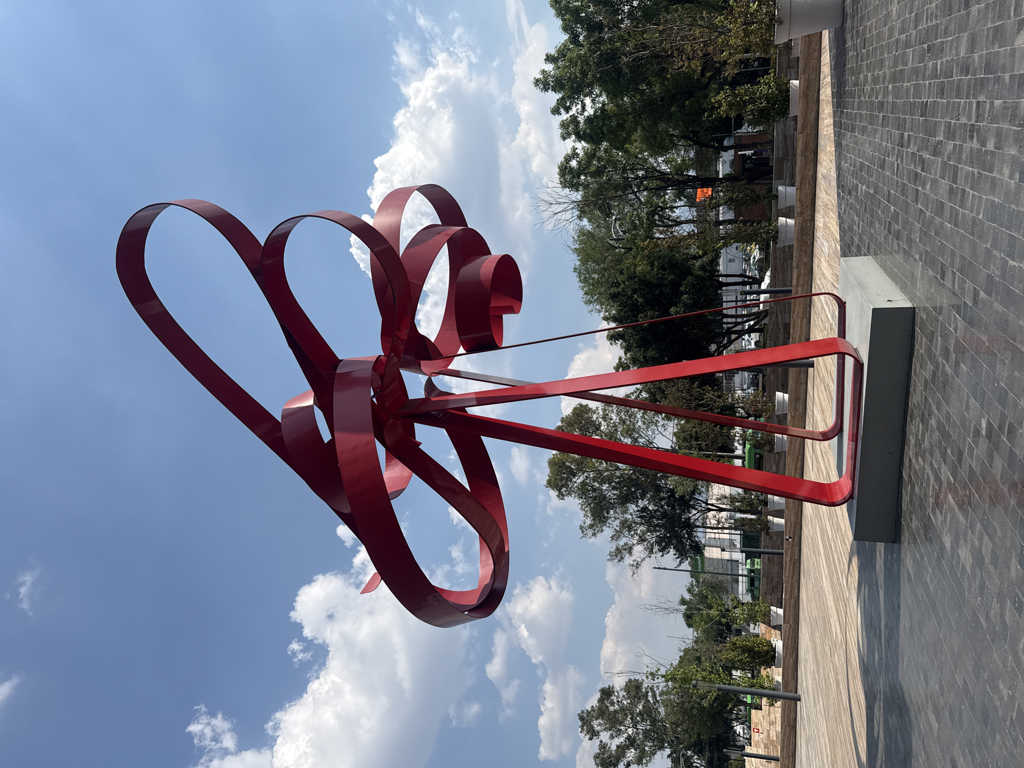 The image shows a large, red metal sculpture of a stylized bow located in an outdoor public space. The sculpture is composed of curved and straight metal bands. The sky is blue with scattered white clouds. Behind the sculpture, there is a row of trees and a paved walkway. The photograph appears to be taken in Mexico City, Mexico.