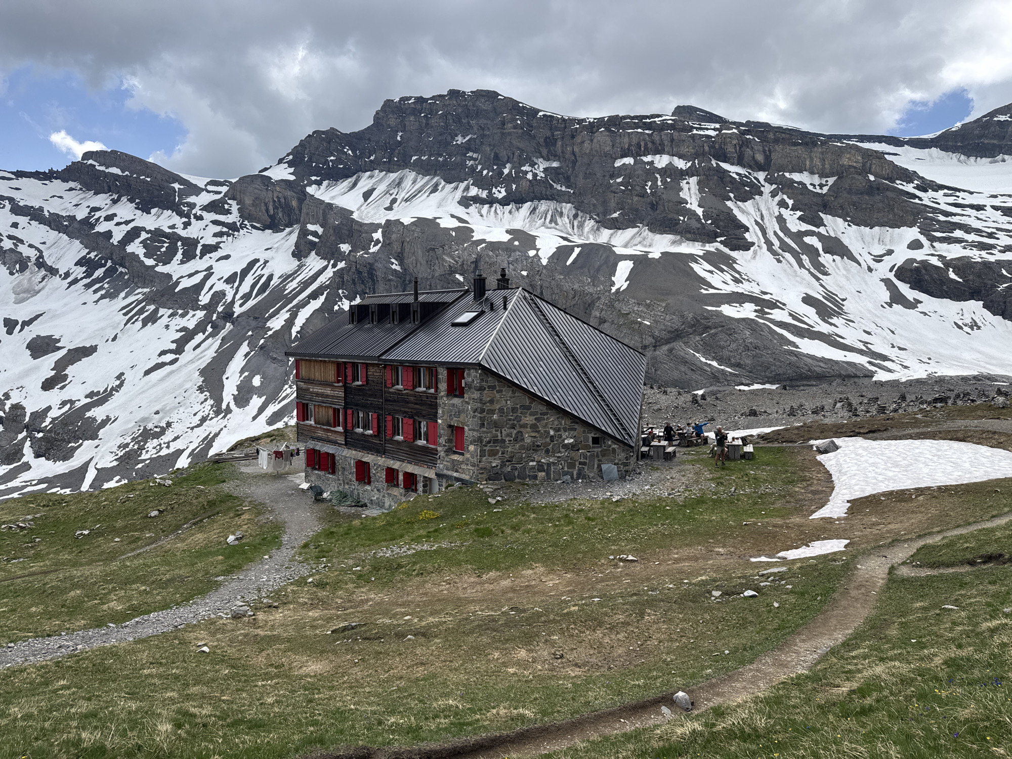 The image shows a stone and wood mountain hut with red shutters, set against a backdrop of snow-covered mountains and a cloudy sky. The hut is located in a grassy, sloped terrain with a gravel path leading towards it. In front of the hut, there are picnic tables with people seated, and a pile of laundry hanging on a line. Further in the background, there are stacks of stones and some patches of snow on the mountainside. The environment suggests a remote, high-altitude location, consistent with the area around Leukerbad, Switzerland, known for its alpine scenery and mountain huts.