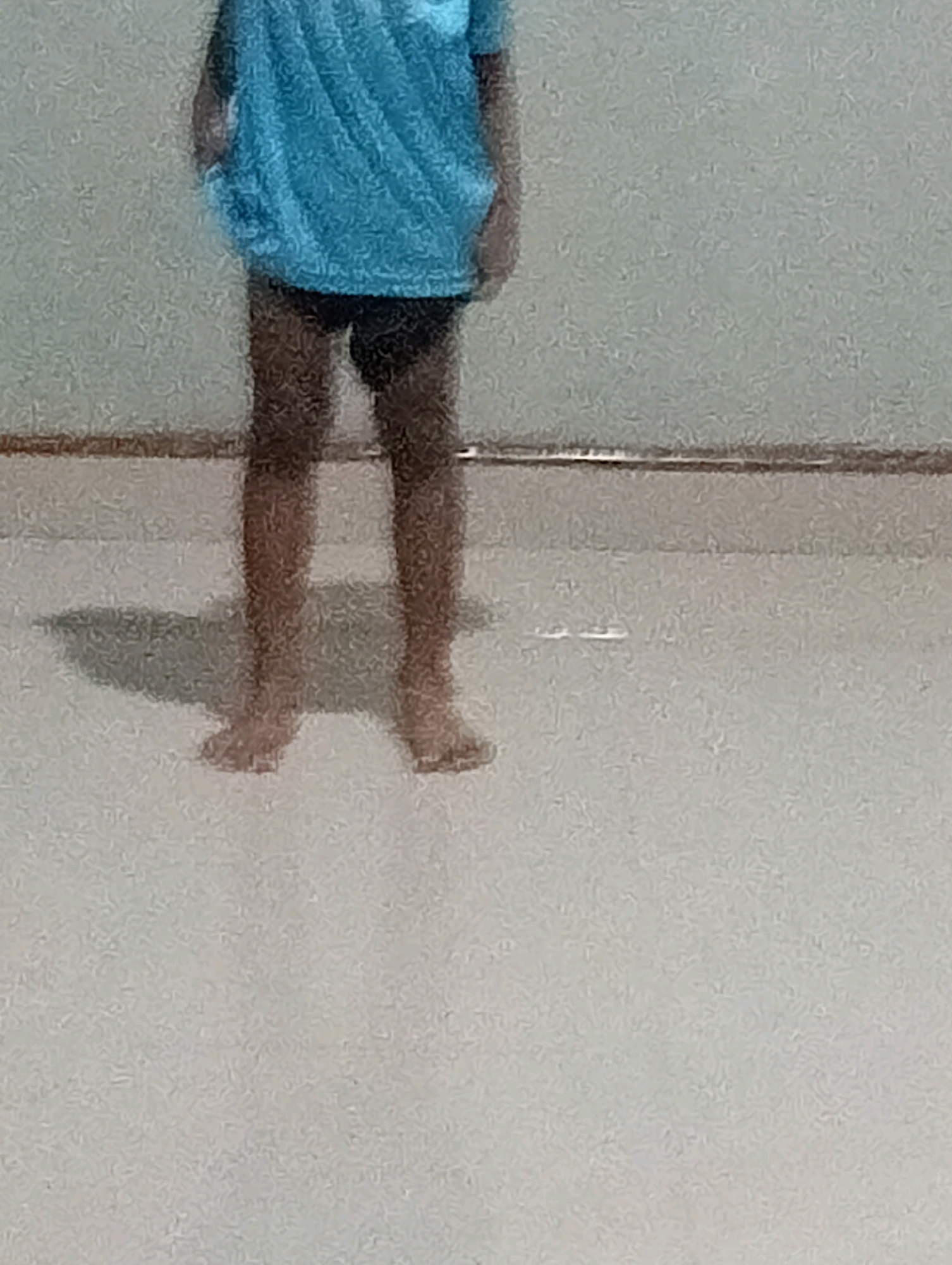 The image shows the lower body of a person standing on a light-colored floor. The person is wearing a blue t-shirt and dark shorts. Their feet are bare. A shadow is cast on the floor to the left of the person's feet. The background appears to be a plain wall with a horizontal line near the bottom. The image is blurry and lacks detail. There is no information present in the image that would allow for its localization to Suka Merindu, Indonesia.