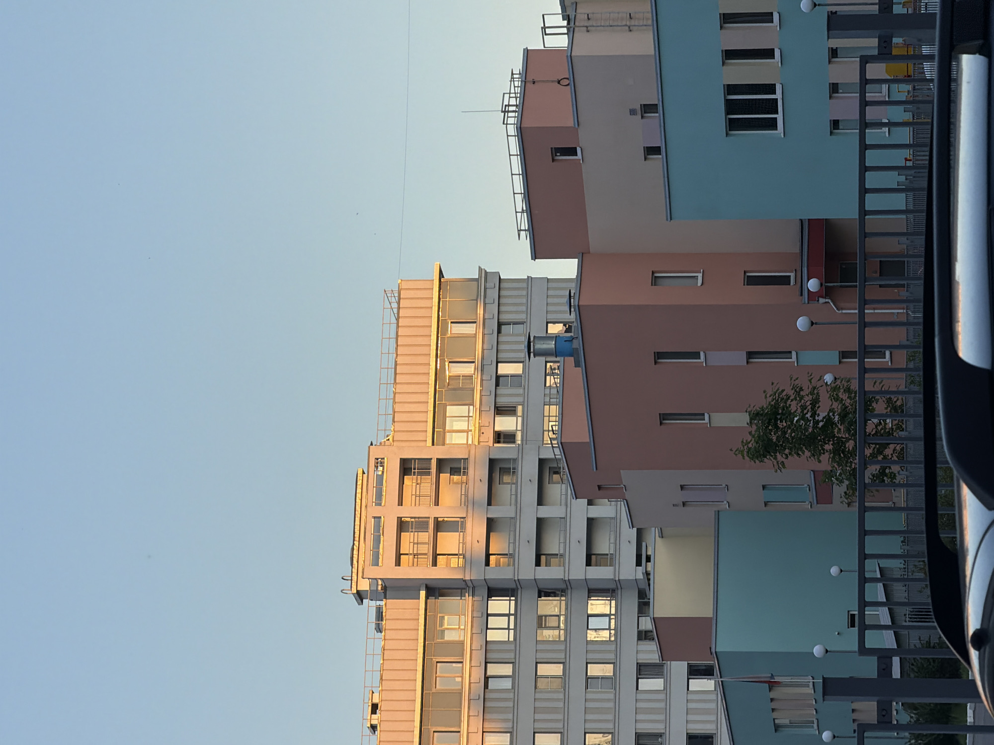 The image presents an urban architectural scene captured during the late afternoon or early evening, evidenced by the warm, golden light illuminating the upper facades of the buildings against a clear, pale blue sky. No individuals are visible within the frame.
In the background, a tall, multi-story residential building stands prominent, characterized by a light grey or beige facade. It features numerous windows, many reflecting the golden sunlight, and multiple staggered balconies. The top section of this building, including its roofline and upper stories, is bathed in direct, low-angle sunlight.
In the mid-ground, a series of smaller, distinct buildings are visible, notable for their varied pastel color palettes, including soft coral-pink, light teal, and pale yellow. These structures exhibit modern, simplified designs with flat roofs. On one of the teal-colored buildings, a flagpole prominently displays a small flag with white, blue, and red horizontal stripes, identifying it as the flag of Russia. Various architectural details are discernible, such as rectangular windows, roof railings, and ventilation units, including a blue cylindrical chimney-like structure on one of the buildings. A small tree with green leaves is positioned among these colorful structures. Several spherical white streetlights are arranged in front of these mid-ground buildings.
The foreground is partially occupied by a dark, metallic railing, likely part of a fence or a vehicle's roof rack. Beneath this railing, the dark, reflective surface of what appears to be the roof of a vehicle, possibly a car or SUV, is visible at the very bottom of the image.
Based on the visible Russian flag and the contemporary architectural styles consistent with modern urban development, the scene is situated in the city of Saint Petersburg, Russia.