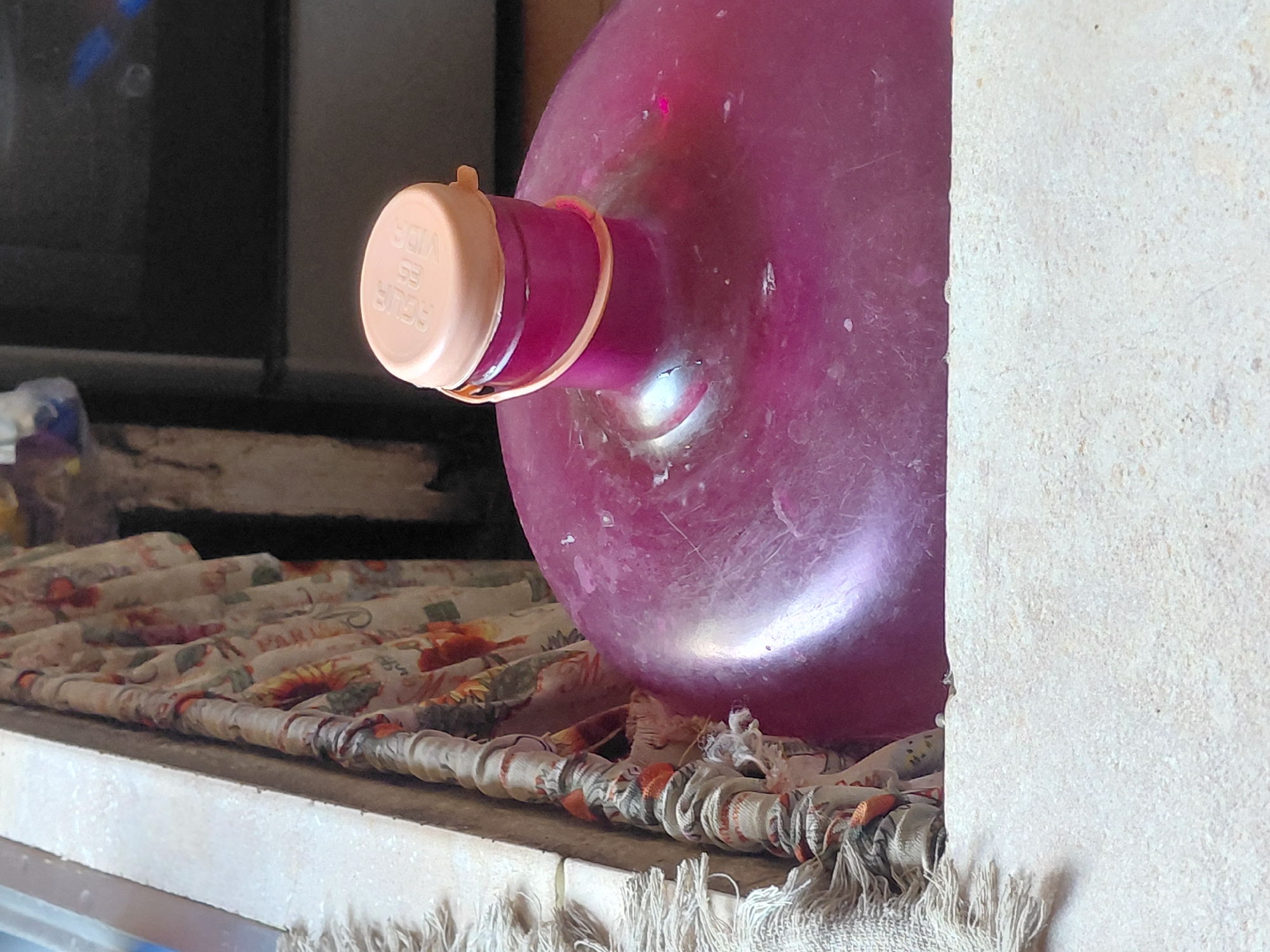 A close-up, eye-level shot shows the top of a large, magenta-colored water jug with a pale pink cap. The jug is positioned on a light-colored surface, possibly a ledge or table, in what appears to be an indoor setting in Santa Ana, Venezuela. The background is blurred, but suggests a domestic environment with a floral fabric hanging on the left and dark cabinetry or appliances on the right. The lid of the jug has some embossed text, partially visible as "AGUA ES VIDA," which translates to "Water is Life." The jug and lid have a slightly scuffed and used appearance. To the left of the jug, a fringed textile, possibly a rug or curtain, is visible. The overall scene is focused on the jug, implying it is the primary subject of interest.