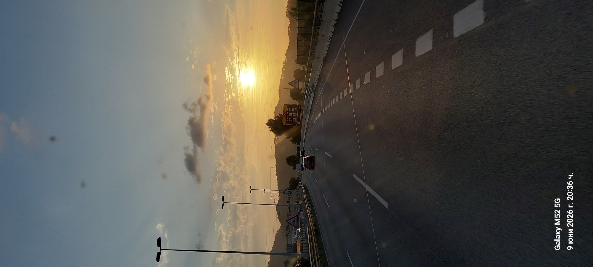 The image captures a highway scene at sunset, likely in or near Castellbisbal, Spain, given the timestamp's reference to "9 юни 2026 г." which is Bulgarian for June 9, 2026. The sun is low in the sky, casting a warm, orange glow over the landscape and creating long shadows. A multi-lane highway stretches into the distance, with several vehicles visible, including cars and what appears to be a van. On the right side of the highway, there is a brown sign with text and icons, including "MONTSERRAT" and an icon that suggests an information point or rest stop. The landscape beyond the highway is hilly, with vegetation and trees, and silhouetted against the bright sky. Tall streetlights are positioned along the side of the road. The sky itself is a gradient of blue and orange, with scattered clouds, and there are some faint circular smudges in the upper part of the image, possibly lens flares or dust on the camera. The bottom of the image displays a watermark indicating "Galaxy M52 5G" and the date and time "9 юни 2026 г. 20:36 ч."