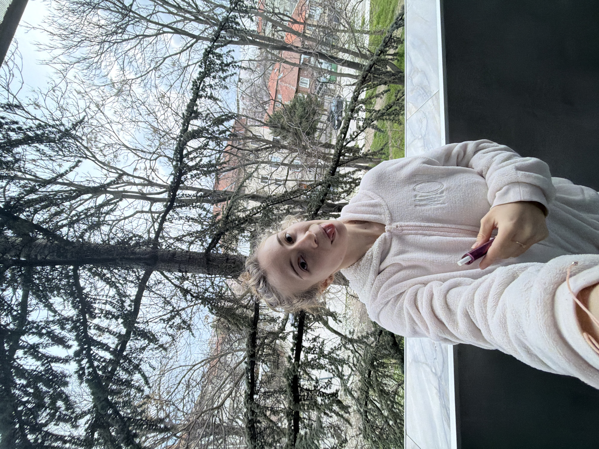 A light-skinned female individual is depicted in a close-up selfie taken from a balcony or terrace overlooking an urban area in Plovdiv, Bulgaria. The subject, positioned centrally, has light-colored hair styled in a bun and wears a light pink, textured, zippered hooded jacket, featuring an embroidered "MILO" on the left chest. She holds a dark pink cylindrical object with a white tip in her right hand. Her left wrist shows a light pink cord. She is looking directly at the camera with a playful expression, her tongue extended. The immediate foreground includes a light-colored, possibly marble-patterned railing, with a dark gray wall section beneath it. The background reveals a dense canopy of trees, primarily bare deciduous trees, interspersed with one prominent evergreen tree. Beyond the trees, characteristic Plovdiv architecture is visible, consisting of multi-story buildings with red-tiled roofs and light-colored facades. A street with a dark-colored vehicle is discernible in the distant background. The sky is overcast.