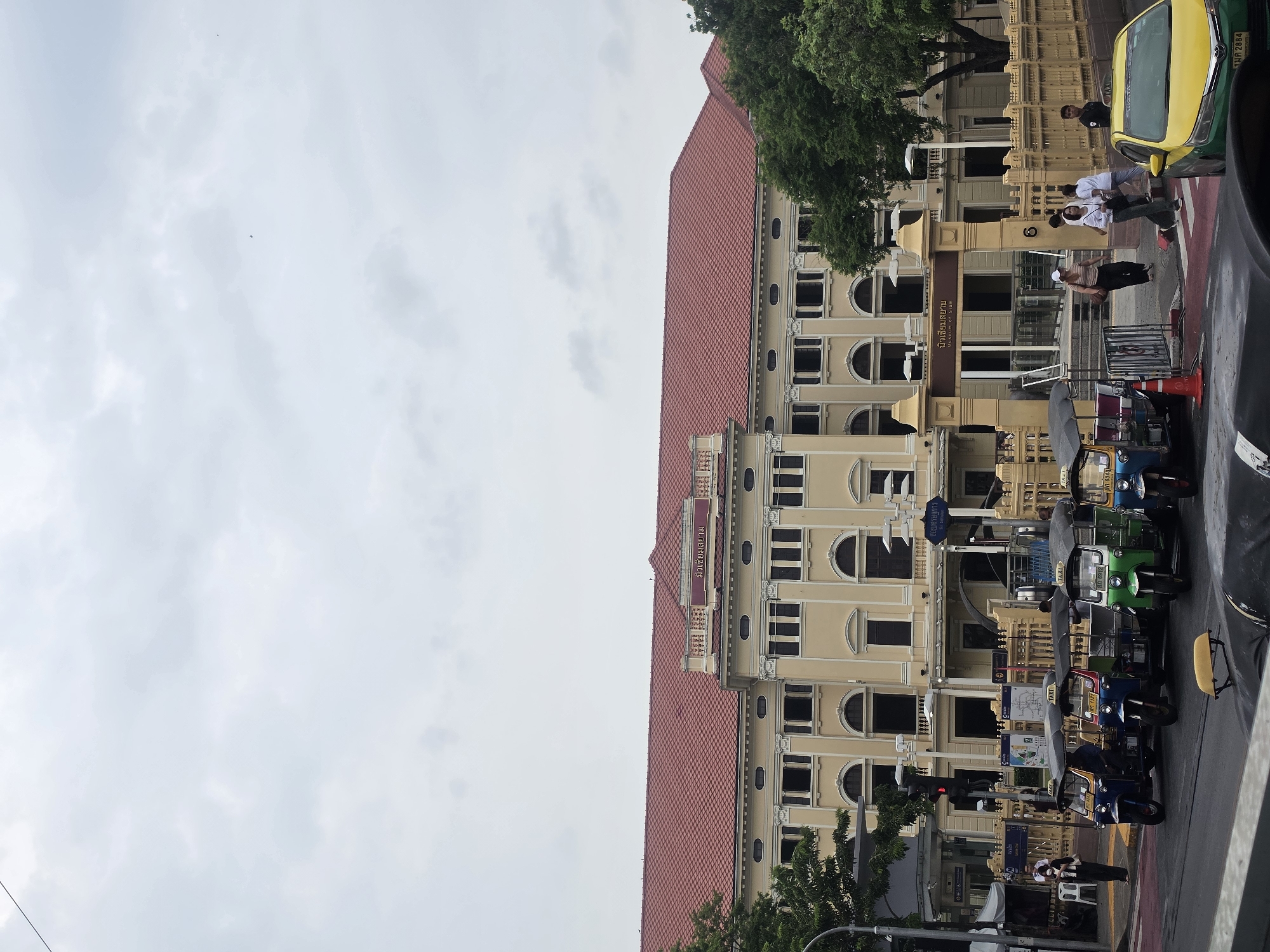 The image shows the Museum of Siam in Bangkok, Thailand. The building has a pale yellow facade with a red-tiled roof. It has multiple arched windows and decorative columns. In front of the building, there are parked tuk-tuks, a yellow and green taxi, and pedestrians on the sidewalk. The sky is overcast. Signs indicating "Museum of Siam" are visible in both Thai and English.