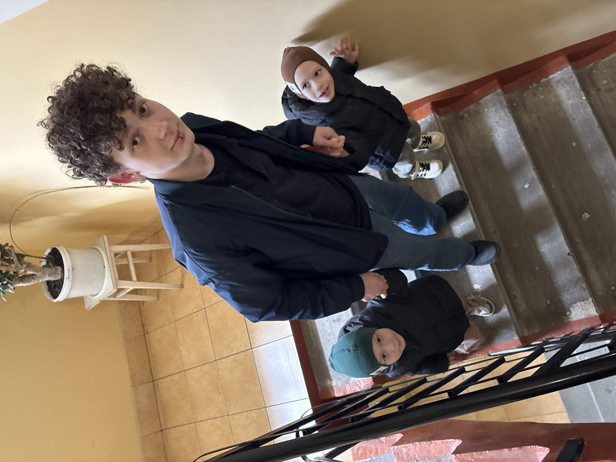 This high-angle image, captured in Ulyanovsk, Russia, features three individuals on a concrete staircase within an indoor setting, likely a residential building. The walls are painted a light yellow or beige, and the landing has a tiled floor with light brown square tiles.
In the foreground, a young man or teenager with curly dark hair, wearing a dark blue jacket and jeans, looks directly at the camera with a neutral expression. Beside him, on the tiled landing, a potted plant sits in a white pot on a small, light-colored wooden stool.
The concrete stairs feature red-painted edges and are bordered by a black metal railing. Further down the stairs, two younger children are visible, both looking up towards the camera. The first child, positioned behind the young man, wears a brown knitted beanie, a black puffy jacket, light-colored pants, and white sneakers with black stripes. This child is smiling brightly and has one hand raised. Below and slightly to the side, a second younger child, wearing a dark green beanie and a black puffy jacket, also looks up with a slight smile.
The lighting in the stairwell is even, without strong shadows, indicating either artificial lighting or an overcast day. The scene captures a casual, posed moment within a common architectural environment found in Russian cities like Ulyanovsk. No visible text is present in the image.