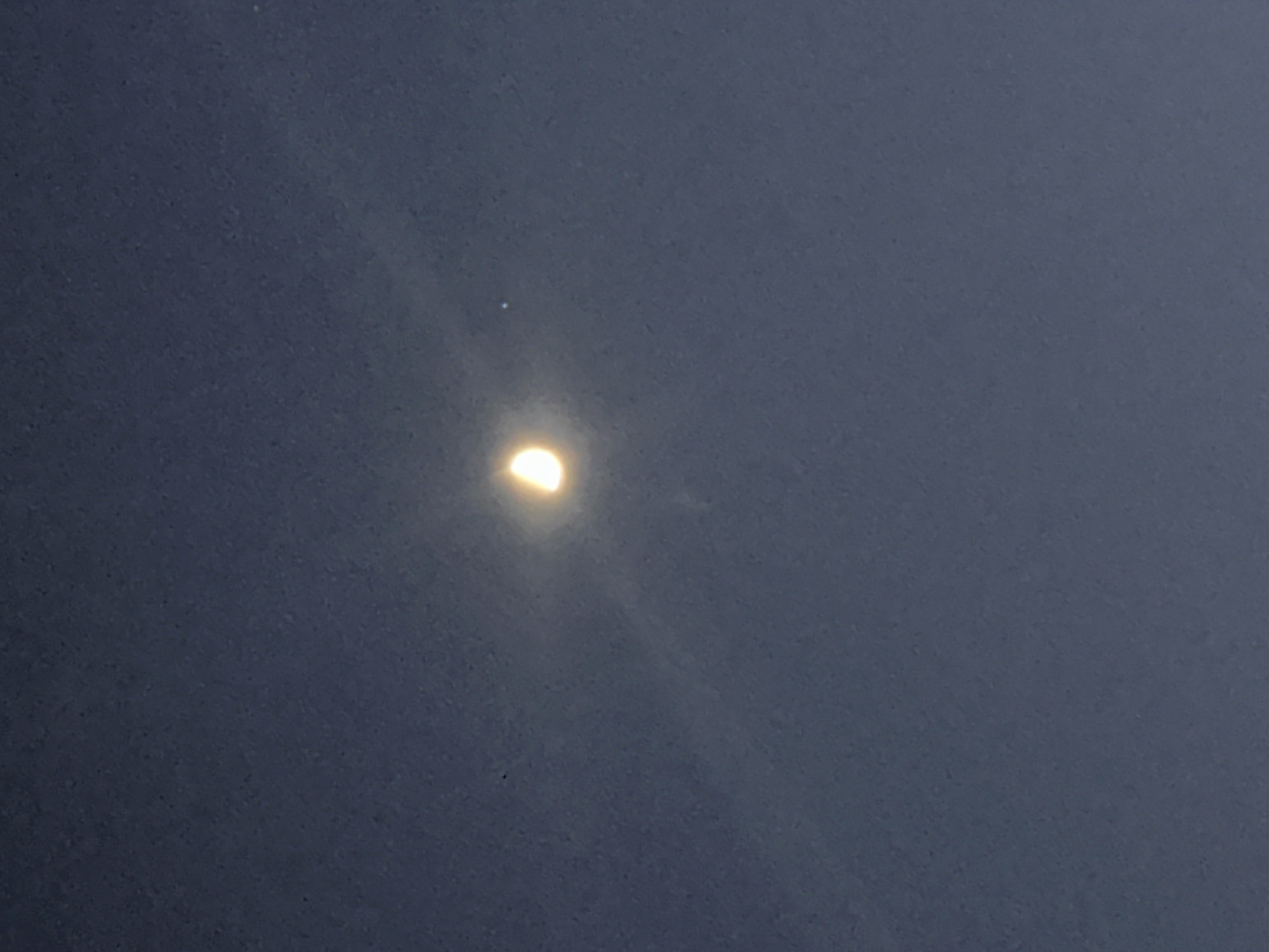 The image is a photograph of the night sky taken in Varna, Bulgaria. The sky is a dark blue, with a faint haziness that diffuses the light. In the center of the frame, the moon is visible, illuminated by about half. A bright halo of light emanates from the moon, creating rays that spread outwards. To the right of the moon, a small, bright point of light, possibly a star or planet, is visible. The overall impression is one of a clear night sky with celestial bodies prominently displayed.