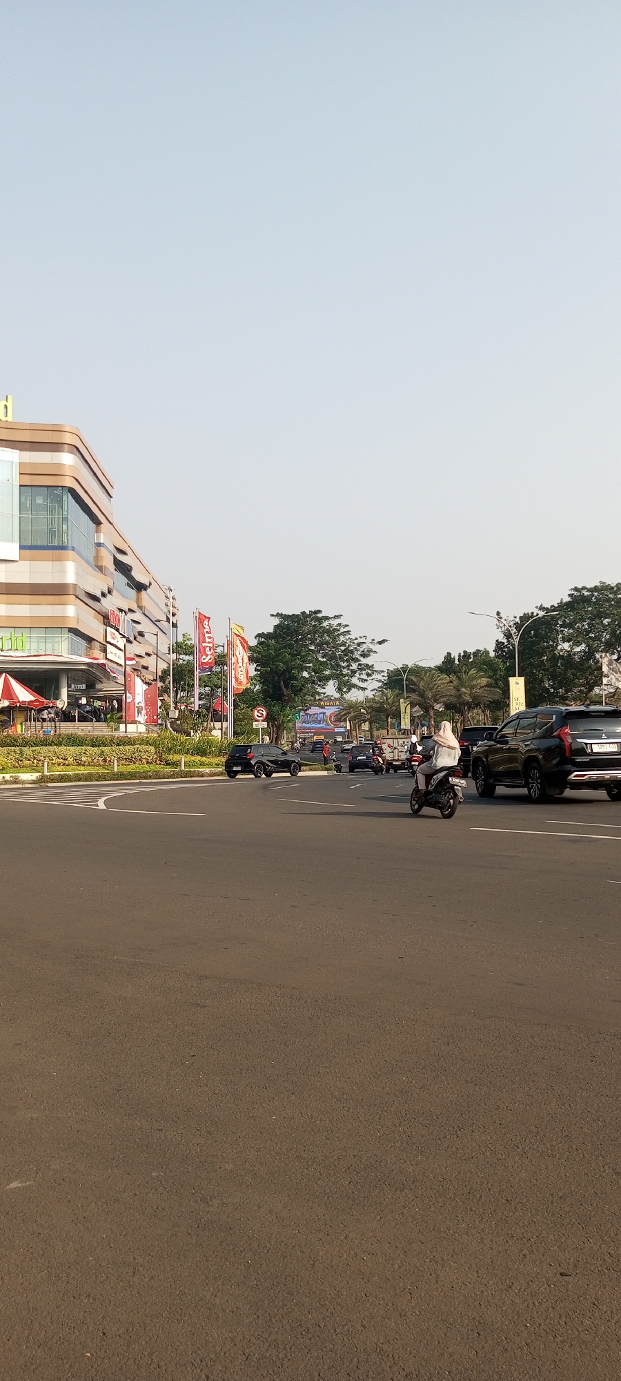 This image captures a street scene in Lambangjaya, Indonesia, during daylight. The foreground is dominated by a wide asphalt road with white lane markings. On the left side of the road, a modern, multi-story building with a beige and metallic facade stands prominently. Red and white flags are displayed in front of the building, suggesting a commercial or event space.
Several vehicles are visible on the road, including a black SUV, a smaller black car, and multiple motorcycles. One motorcycle, with a rider wearing a light-colored hijab, is in the center of the frame, moving forward. Traffic appears to be moderate.
The background features a line of trees, including palm trees, and streetlights. The sky is clear and light blue, indicating a sunny day. The overall impression is that of a busy urban environment with commercial activity.