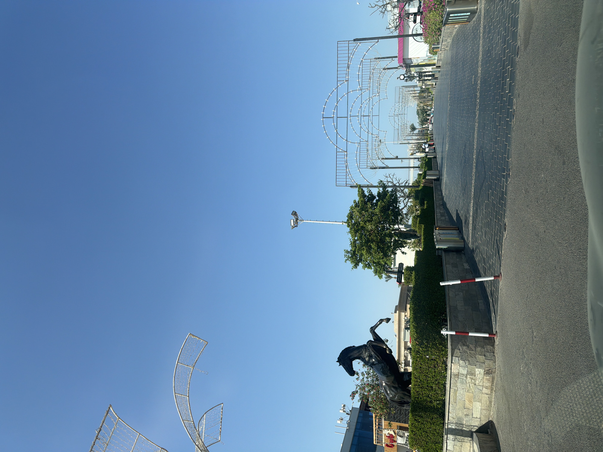 The image depicts an outdoor scene in Sharm El Sheikh, Egypt, likely a public thoroughfare or entrance to a commercial area. The foreground shows a paved road with a textured surface, possibly cobblestone or interlocking pavers, leading into the distance. On the left, a large, dark statue of a rearing horse stands prominently, positioned atop a well-maintained green hedge that is bordered by a stone wall. Behind the horse statue, a modern building with blue glass windows is partially visible.
The pathway is flanked by decorative arched structures adorned with white lights, creating an illuminated pathway. These arches recede into the background, suggesting a long, grand entrance or promenade. Several trees are scattered throughout the scene, including a large, leafy tree to the right of the horse statue and other trees and palm trees further down the path. The sky is a clear, bright blue, indicating a sunny day.
In the distance, the pathway appears to lead towards a collection of buildings and potentially more decorative structures. A few individuals are faintly visible walking along the path in the mid-ground, suggesting moderate pedestrian activity. Red and white striped bollards are present near the road's edge, likely for traffic control or demarcation. The overall impression is one of an attractive, possibly tourist-oriented, urban or commercial space designed with decorative elements.