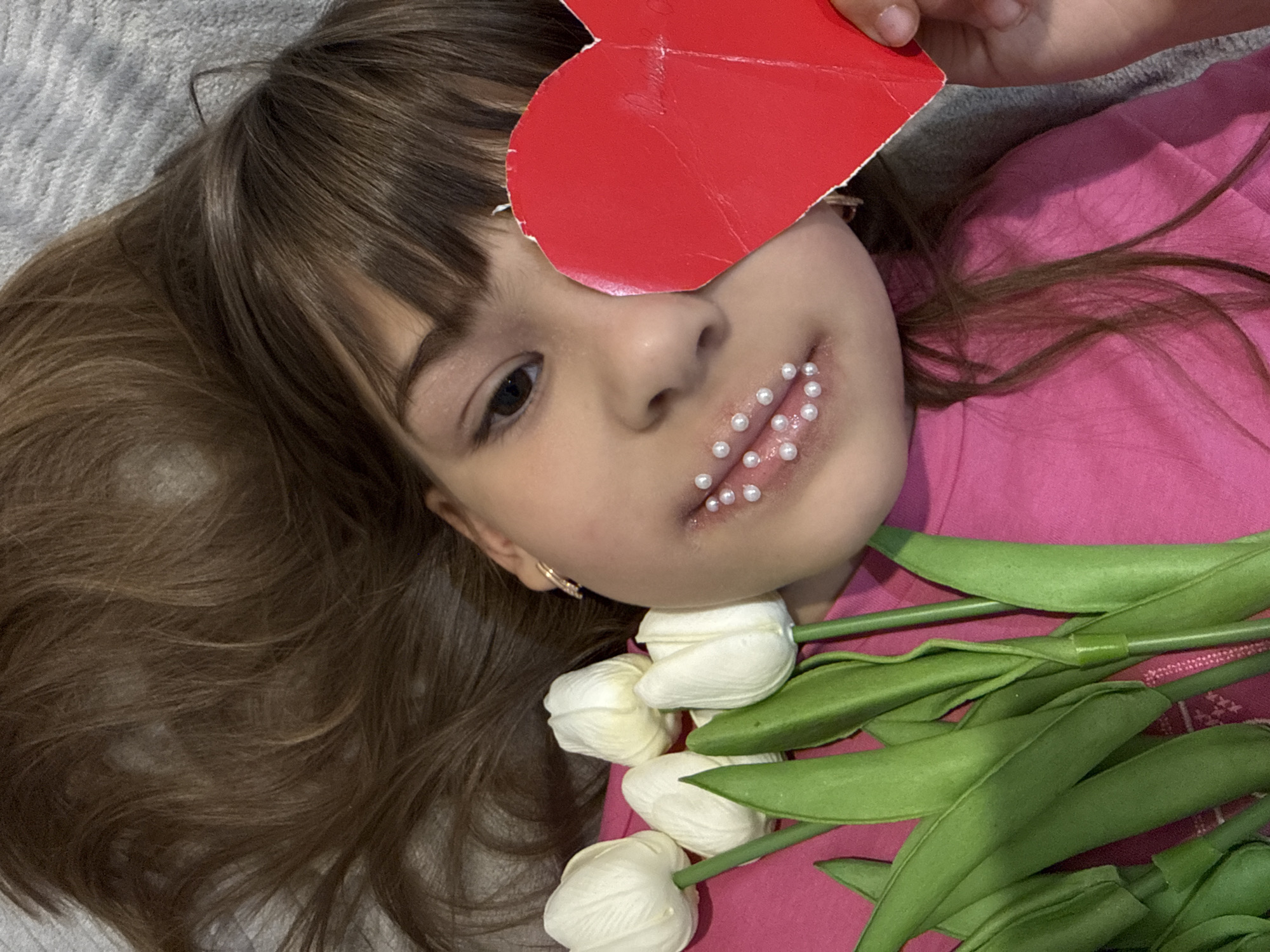 A young girl with long, brown hair lies on her back, looking upwards towards the camera. Her head is slightly tilted, and her expression is neutral, with her visible left eye open. A red, somewhat crudely cut heart shape made of paper or card is held by a hand above her right eye, covering it. Her lips are adorned with numerous small, white pearls, arranged in decorative clusters on both the upper and lower lip. She is wearing a bright pink top.
To her right, a bouquet of several white tulips with green stems and leaves rests beside her face and on her pink top. The background consists of a soft, textured grey surface on the left, likely a blanket or rug, and her pink clothing. The lighting appears soft and even, suggesting an indoor setting. No text is visible in the image.