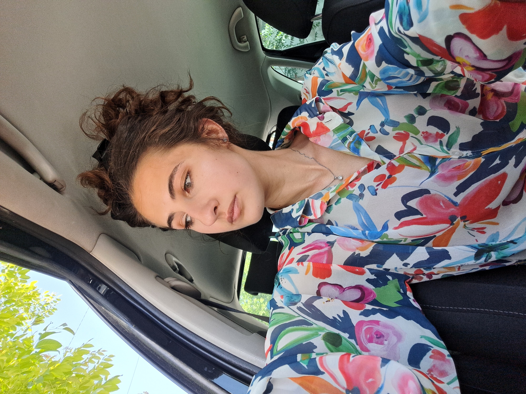 A young woman is sitting inside a car. She has fair skin, brown curly hair pulled back in a bun, and wears a floral-patterned blouse. She is looking downwards with a pensive expression. The interior of the car is visible, including the headrest of the seat, the car door, and part of the dashboard. Through the car window, there are green trees and a hint of blue sky. The location is probably Sofia, Bulgaria, based on the license plates on cars in similar photographs and the presence of vegetation found in the urban areas of Sofia. The photo appears to have been taken at an upward angle.