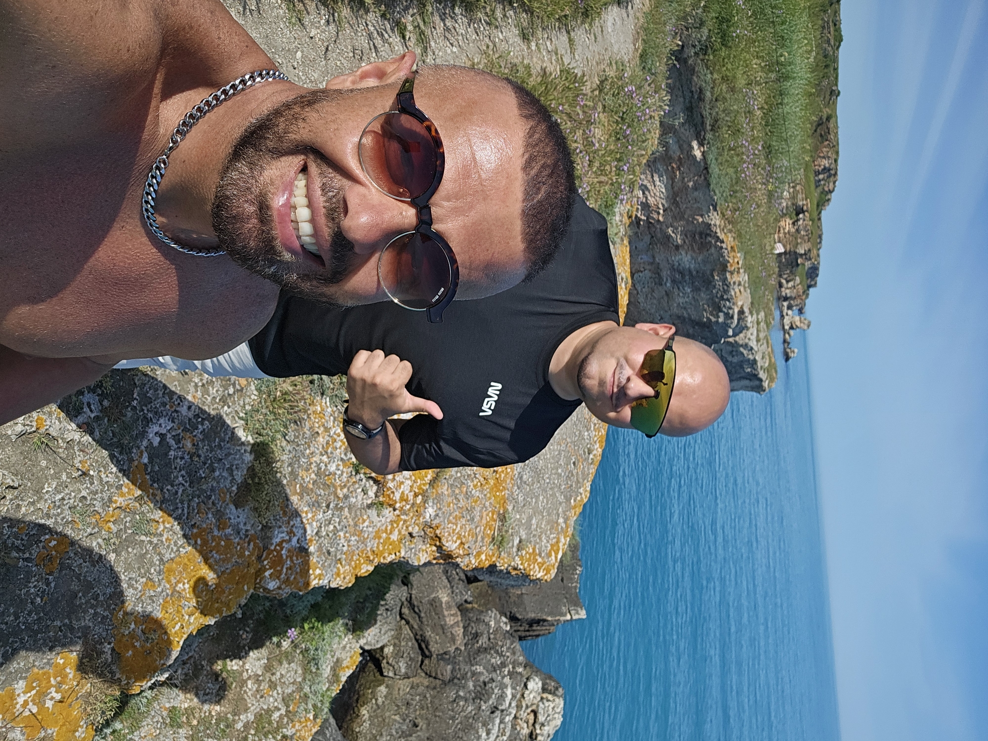 Two bald men are standing on a cliff. The man on the left is shirtless and wearing sunglasses and a silver chain. He is smiling. The man on the right is wearing a black t-shirt, sunglasses, and a watch. He is pointing with his left hand. They are standing on rocks with some vegetation, overlooking the ocean. The location is Kamen bryag, Bulgaria.