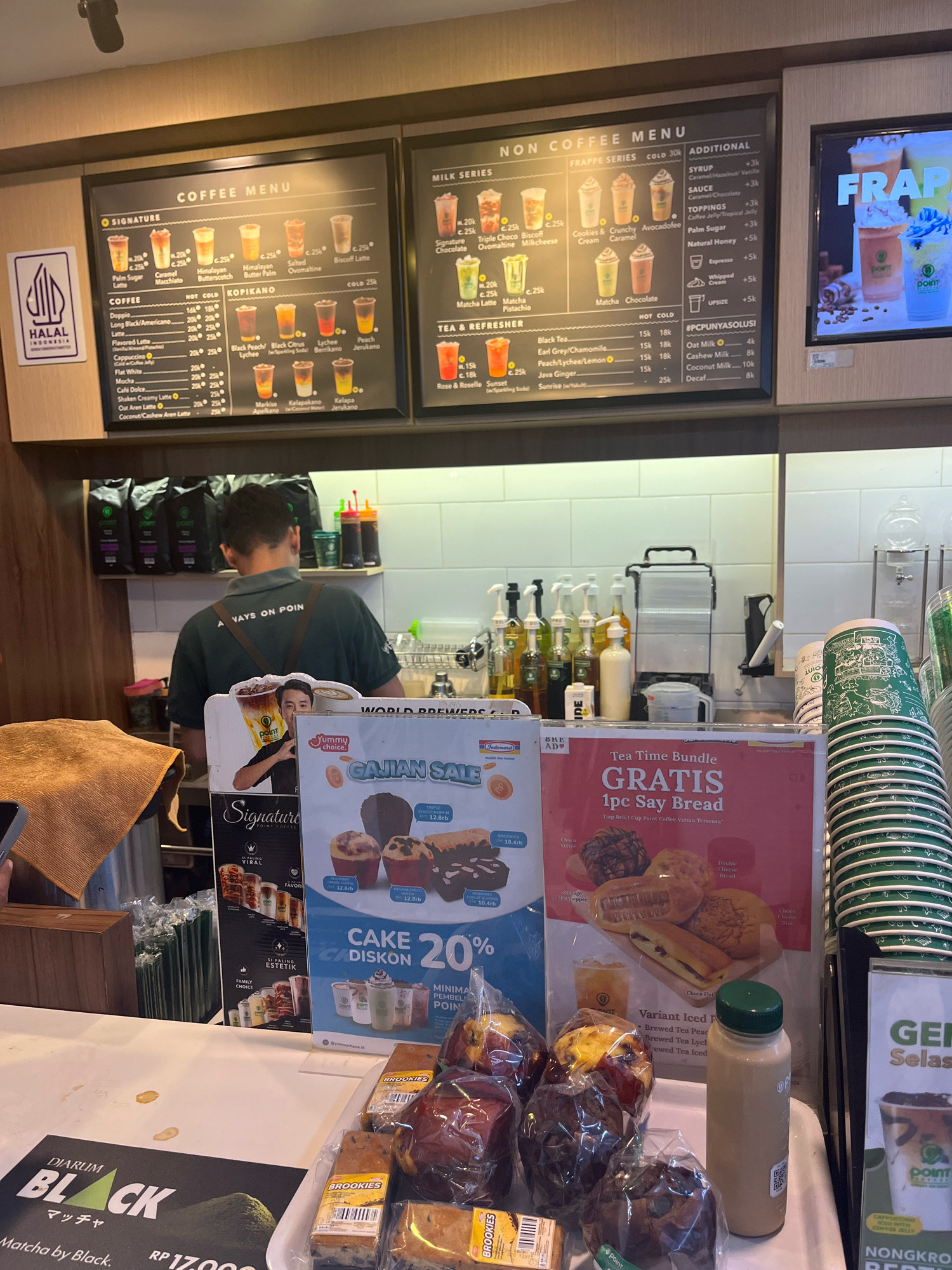 The image depicts the interior of a coffee shop in Banguntapan, Indonesia. A male barista, wearing a dark green t-shirt with the text "Aways on Point" on the back and an apron, is seen working behind a counter. The counter is cluttered with various items including coffee beans in bags, syrup bottles, cups, a coffee machine, and packaged pastries and bread.
Dominating the background is a large menu board displaying "COFFEE MENU" and "NON COFFEE MENU" with various drink options and prices. To the right, a digital screen shows a colorful advertisement for "FRAPPE". There are also several promotional signs on the counter, including one for a "Tea Time Bundle" offering a free "Say Bread" with a cup of Point Coffee. Another sign promotes a "CAKE DISKON 20%".
In the foreground, packaged pastries and loaves of bread are displayed on the counter. A bottle of iced coffee and a stack of green paper cups are also visible. A sign on the far left of the image displays "DIARUM BLACK MATCHAA" with an image of an avocado. The overall environment suggests a busy coffee shop setting.