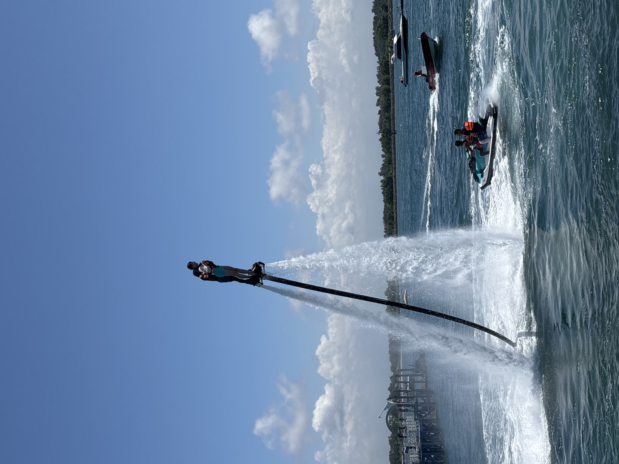 A person is using a flyboard over the water near Tanjung Benoa, Indonesia. The flyboard is connected to a hose that is pumping water to propel the person into the air. A jet ski with two people is nearby, and another boat is further away. The sky is blue with some clouds. The water is choppy. A structure is also visible on the shore.