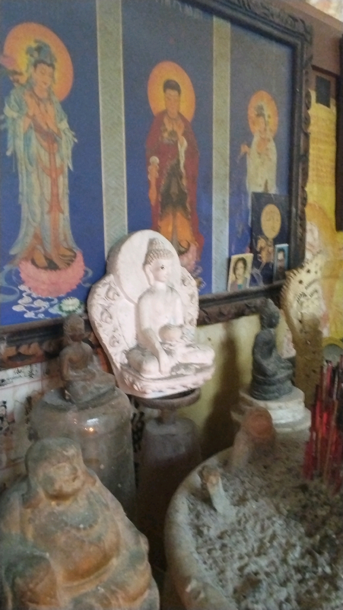A framed painting of three figures, possibly deities, is displayed on a wall. To the left, a figure in flowing blue and white robes holds hands in prayer, with an orange halo. In the center, a figure in a red robe, also with an orange halo, appears to be holding something. To the right, a figure in white, with an orange halo, stands with hands clasped. Below the painting, several Buddha statues are arranged. A large white statue of Buddha in a seated position with intricate carvings is prominent in the foreground. To its left and slightly behind, a smaller, dark bronze Buddha statue is seated. In the lower left of the image, a large, round, stone Buddha statue is partially visible. To the right of the white Buddha statue, a dark stone Buddha statue is seated on a pedestal. In the far right, a bundle of red incense sticks is visible. The overall setting appears to be an indoor space, possibly a temple or shrine, with a dimly lit atmosphere. The specific location in Penompo, Indonesia, is not visually evident from the image itself.
