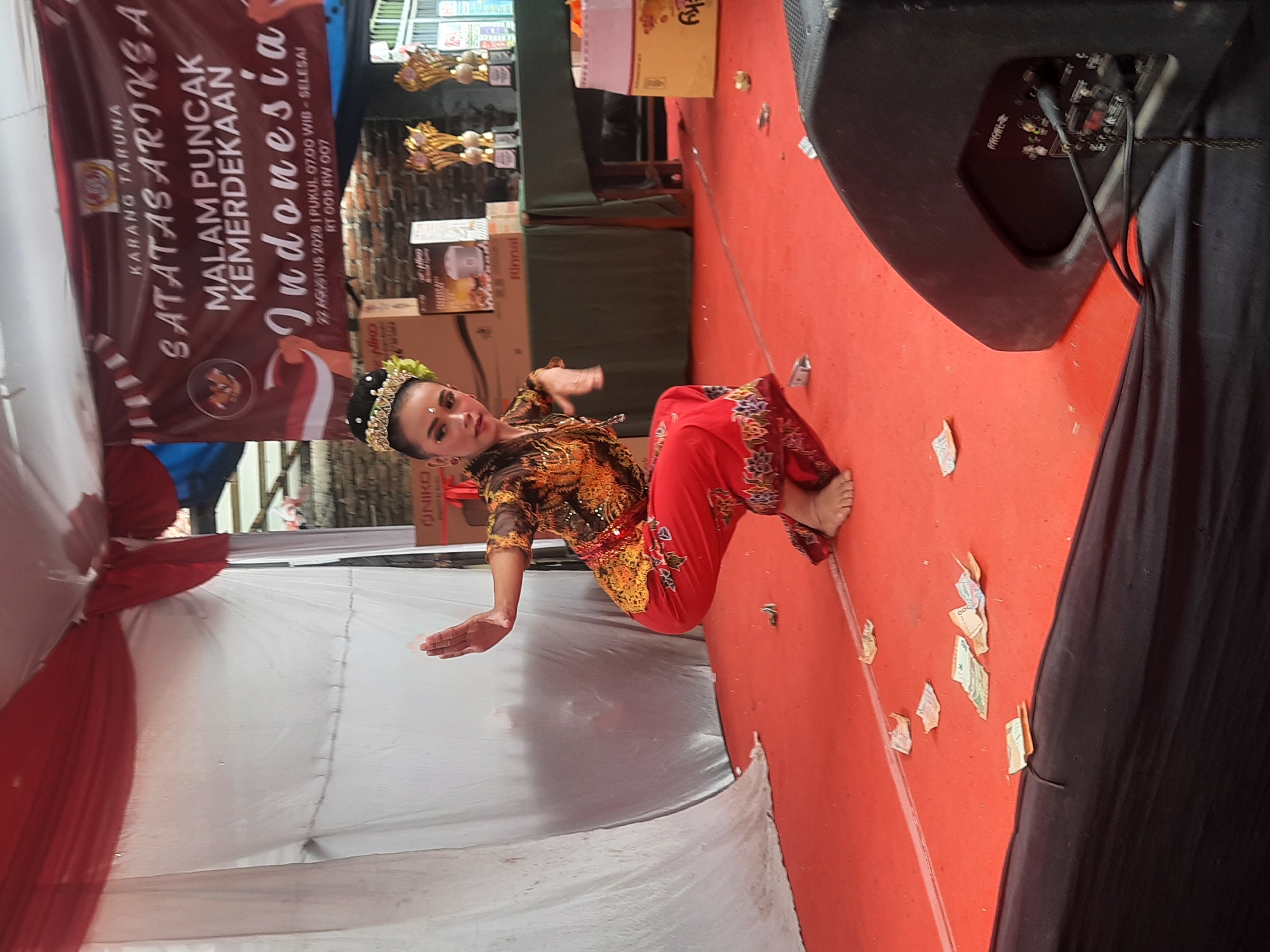 A woman in traditional Indonesian attire, including a bejeweled headpiece, a gold and brown patterned top, and red pants with a colorful hem, performs a dance on an orange stage. Her left arm is extended outward, and her right hand is bent at the elbow. The stage is decorated with a red and white banner in the background that reads "MALAM PUNCAK KEMERDEKAAN INDONESIA" and the date "22 AGUSTUS 2025". There are several stacks of money scattered on the stage floor. To the right of the stage, there is a large black speaker. The event appears to be a celebration of Indonesian Independence. No specific location in Bandung City, Indonesia is discernible from the image.