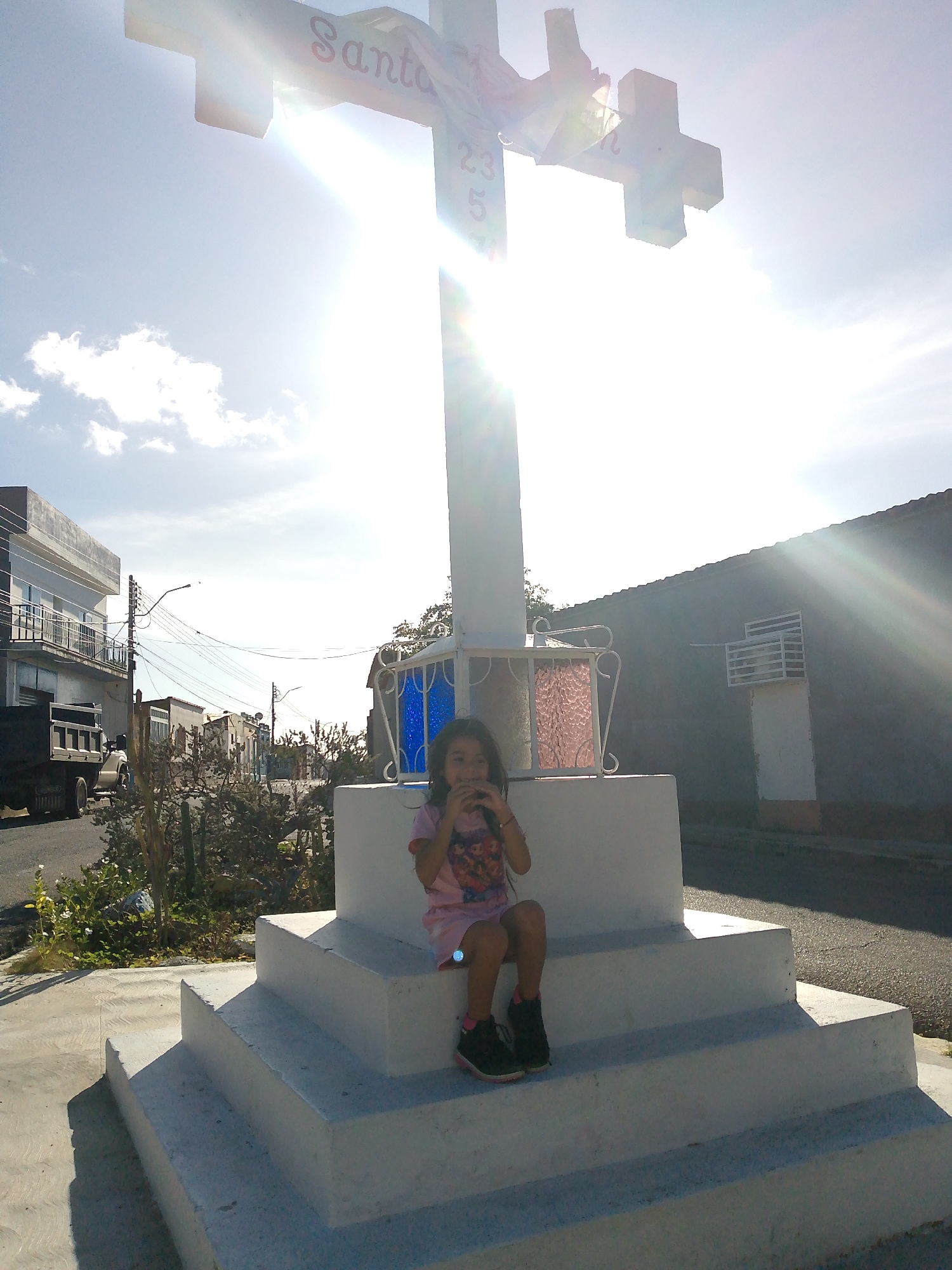 The image depicts an outdoor daytime scene in Juan Griego, Venezuela, centered on a large white concrete cross monument. A young female child is seated on the second step of the monument's three-tiered base. She wears a pink short-sleeved top with a graphic, light-colored shorts or skirt, and dark sneakers with pink accents. Her hands are raised to her mouth, holding an object.
The cross monument features red inscriptions, including "Santa" on its horizontal beam and "23 5" on its vertical beam. Its base incorporates decorative white wrought iron panels containing blue and pink/red glass inserts.
The sun, positioned directly behind the cross, generates a strong lens flare across the upper right quadrant of the image, contributing to the bright, partially cloudy sky. In the background, a paved street runs parallel to the monument. To the left, a multi-story building with grey exterior walls and white balconies is visible, alongside a dark-colored truck and a utility pole with overhead wires. To the right, a single-story building with a dark, textured facade and a white grated fixture is present. Sparse vegetation, including cacti-like plants, lines the street. Shadows cast by the cross monument are visible on the ground in the foreground.