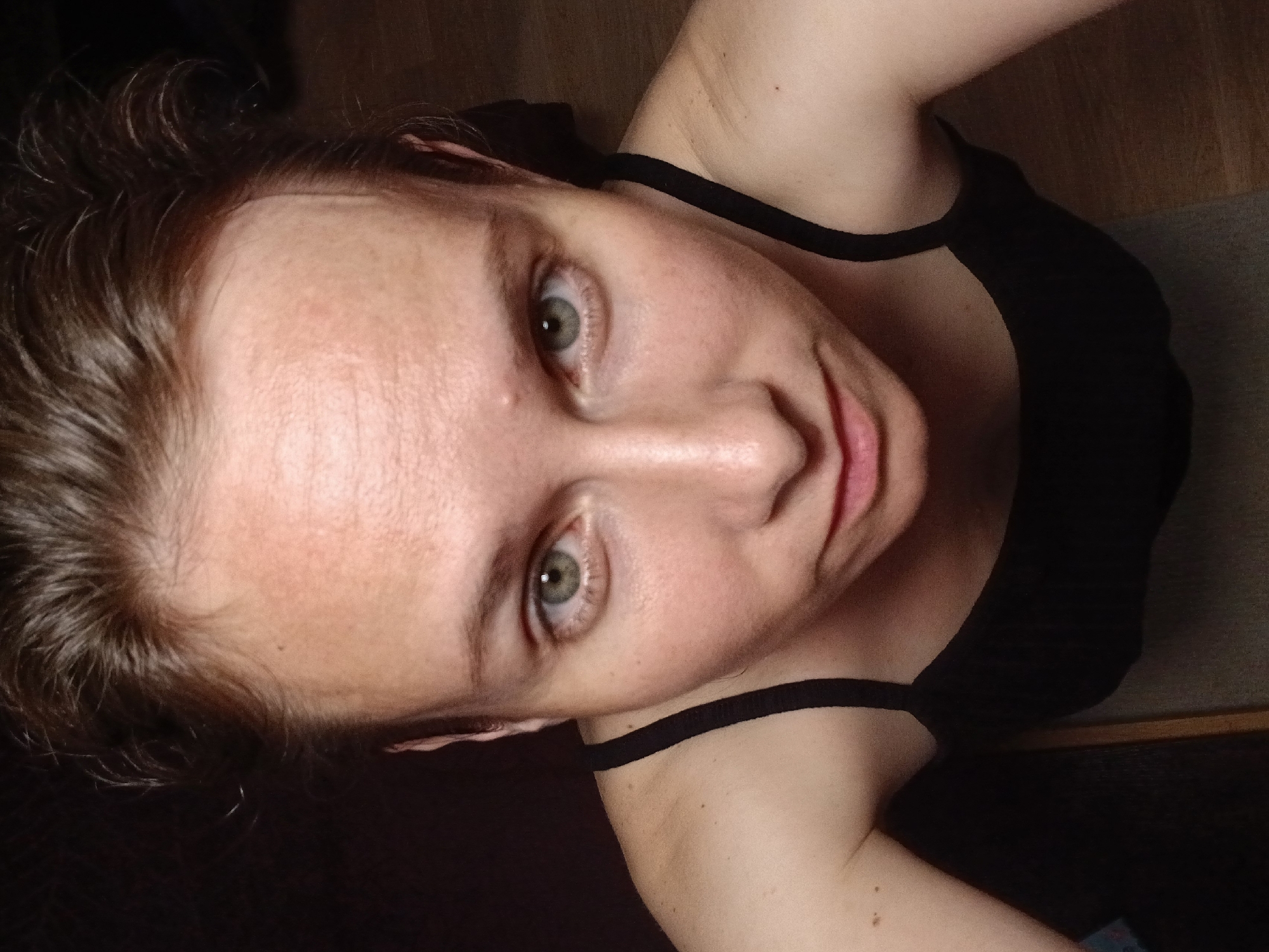 A woman with fair skin, light brown hair, and green eyes looks directly into the camera. She is wearing a black tank top. The image appears to be a selfie taken from a high angle. The background is dark and indistinct, with hints of wooden flooring. There is no information in the image to suggest a location in Yekaterinburg, Russia.