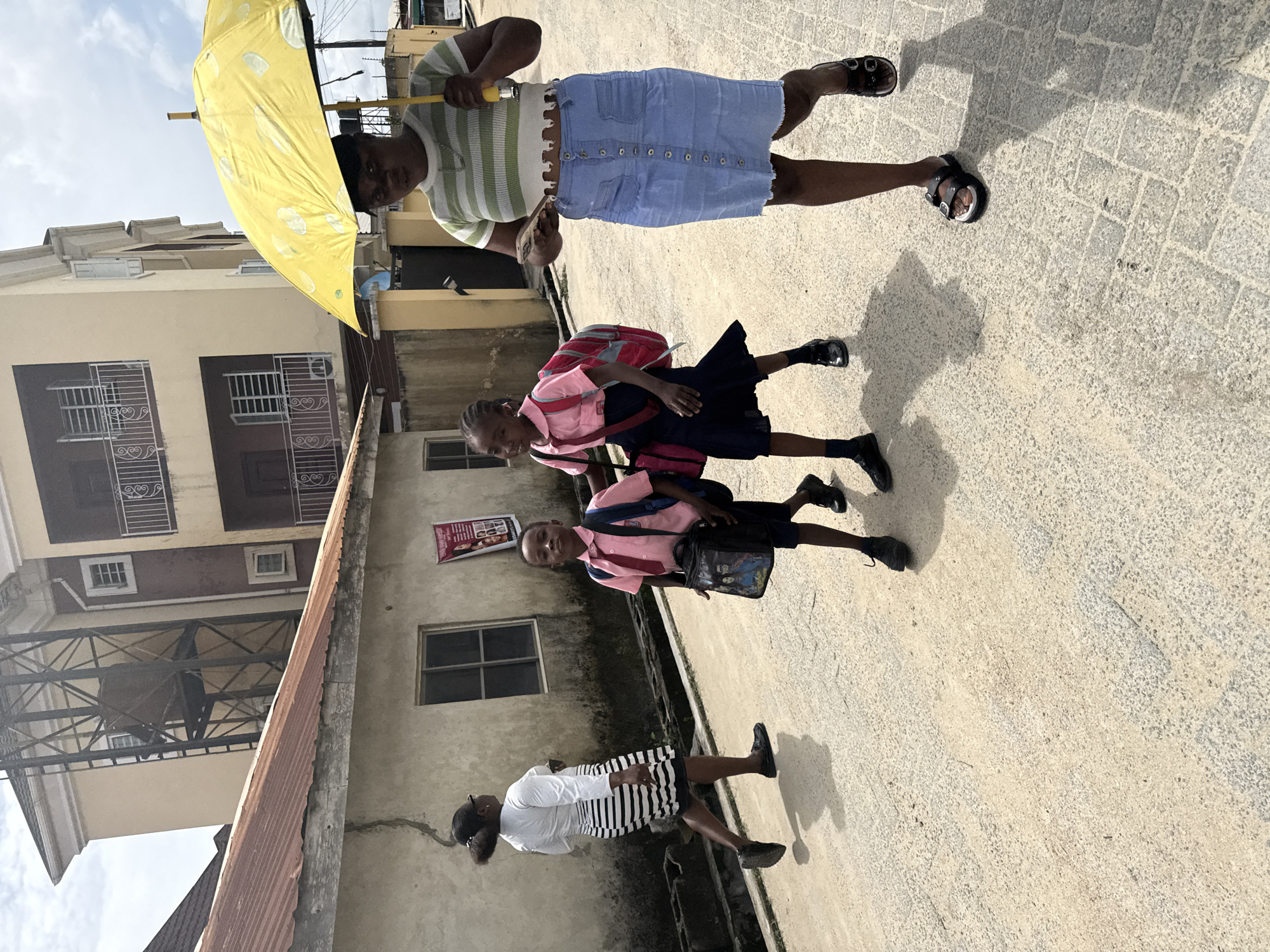 This image captures a bright daytime street scene in Ado, Nigeria, featuring five individuals walking along a paved path bordered by multi-story buildings.
In the foreground, a woman walks holding a bright yellow umbrella with white polka dots, likely for sun protection. She is dressed in a light green and white striped short-sleeved top with a ruffled hem, a light blue denim midi skirt with a front button-up closure, and black sandals. She holds a mobile phone in her left hand. Accompanying her are two children, likely school pupils, dressed in matching pink short-sleeved shirts, dark skirts or shorts, and black shoes. One child carries a red backpack, while the other carries a black shoulder bag.
Further back on the pathway, another woman is visible, wearing a white long-sleeved top and a black and white horizontal striped skirt, with black shoes. The ground beneath them is a light-colored, textured concrete or paved surface, marked by distinct shadows from the people, indicating strong overhead sunlight.
The background consists of several multi-story buildings, characteristic of an urban or suburban environment. These structures feature painted walls, windows often fitted with security bars, and balconies with metal railings. A section of a corrugated metal roof or awning is also visible. Above the buildings, a clear, bright sky can be seen. On one building's wall, a red poster is affixed, which appears to be an advertisement, featuring images of people and some text, with the word "BEAUTY" possibly discernible. The scene depicts a daily routine in a bustling community under sunny conditions.