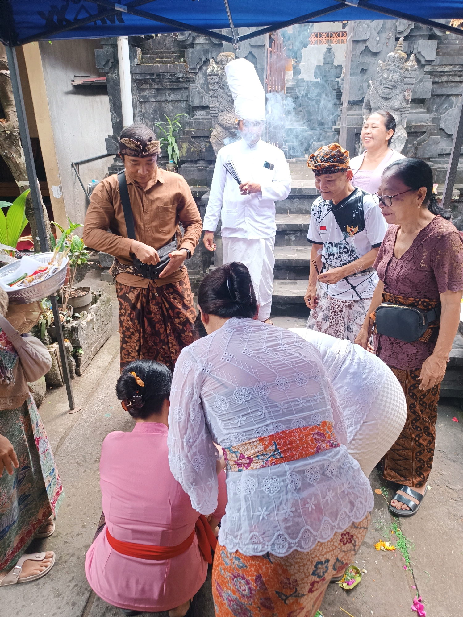 A group of people are gathered outdoors, possibly for a religious ceremony, in Tabanan, Indonesia. In the foreground, a person is kneeling, wearing a pink top and a red sash around their waist. To their right, another person is also kneeling and wearing a white lace top with an orange and floral patterned sarong. A man in a brown long-sleeved shirt and patterned sarong stands to the left, holding a dark bag. To his right, a man in a white outfit with a tall hat is holding incense sticks, with smoke rising. Behind him, a smiling woman in a light pink top is standing, and to her right, a woman in a brown lace top and patterned sarong, wearing glasses and a dark fanny pack, is looking to the side. In the background, there are stone stairs and intricate stone carvings, suggesting a temple or traditional Balinese architecture. The ground is paved and there are some decorative elements and plants present.