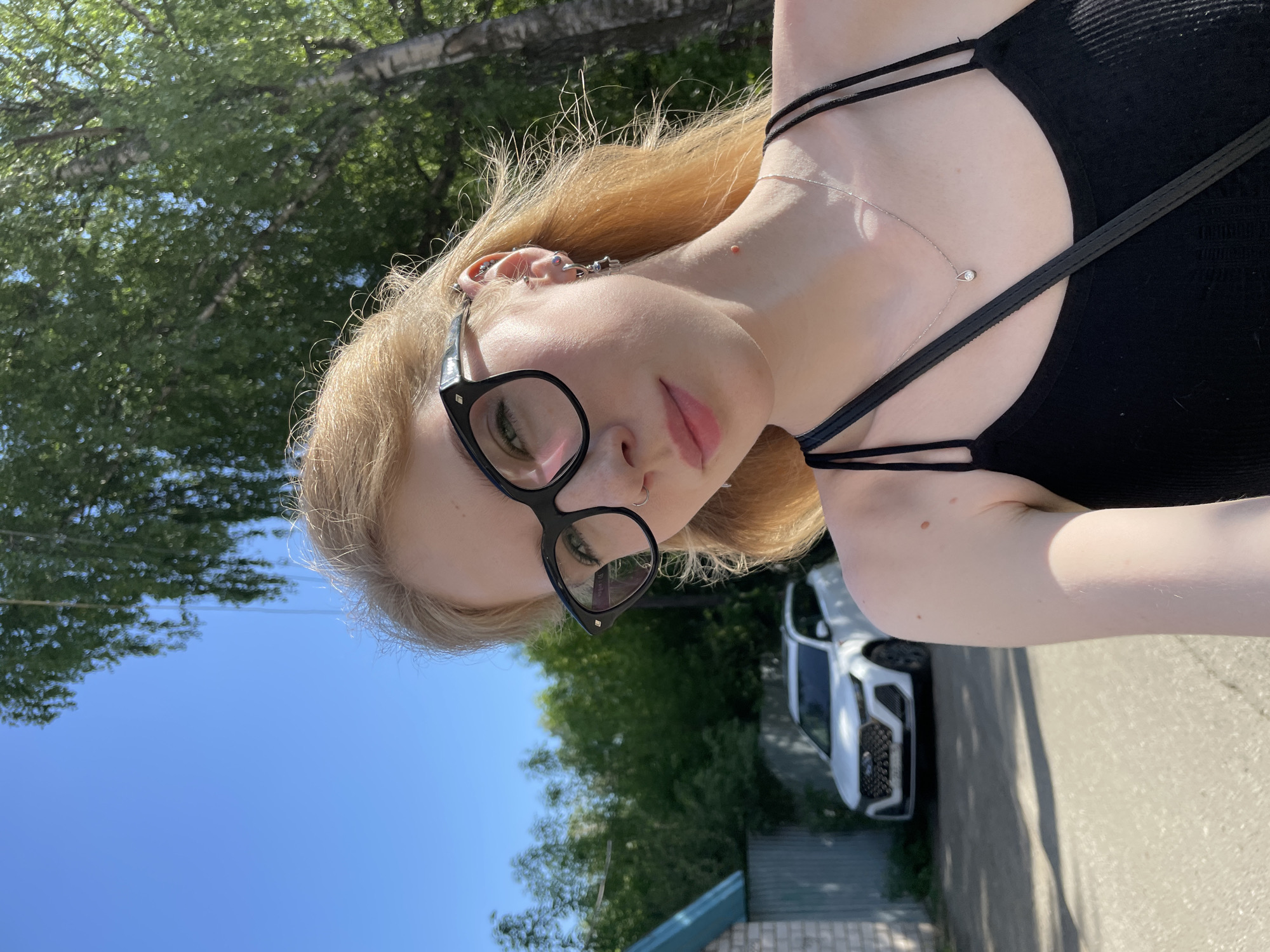 A woman with blonde hair, wearing glasses, a black top, and a necklace, is posing for a photo outdoors. The background shows a blue sky, trees, a building with a blue roof, and a white car. The location appears to be in Izhevsk, Russia.
