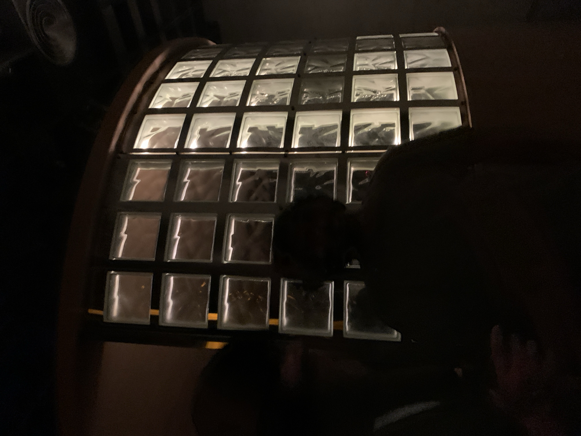 The image shows a curved wall made of glass blocks. The wall appears to be lit from behind, making the blocks glow. A dark, silhouetted figure is partially visible in the foreground, obscuring some of the glass block wall. The location is unknown and no details from the city of Tel-Aviv, Israel are visible.