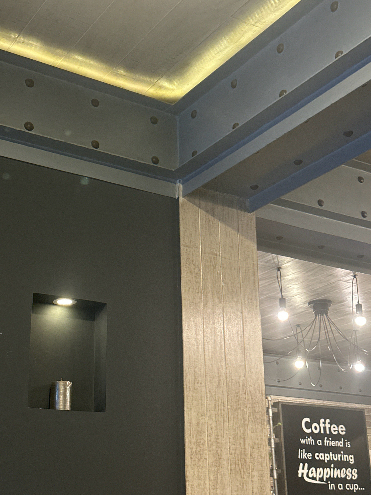 The image captures an interior view of what appears to be a coffee shop or restaurant in Kathmandu Metropolitan City, Nepal. The scene is dominated by architectural elements, including industrial-style exposed beams and a textured ceiling with integrated lighting. On the left, a dark gray wall features a recessed niche illuminated by a spotlight, containing a metallic cylindrical object that could be a candle or a decorative item. To the right, a pillar with a light wood-grain texture stands prominently. In the background, a cluster of exposed light bulbs hangs from the ceiling, creating a warm, ambient glow. A portion of a black sign with white text is visible on the right, featuring a quote about coffee and happiness. No people are visible in the frame.