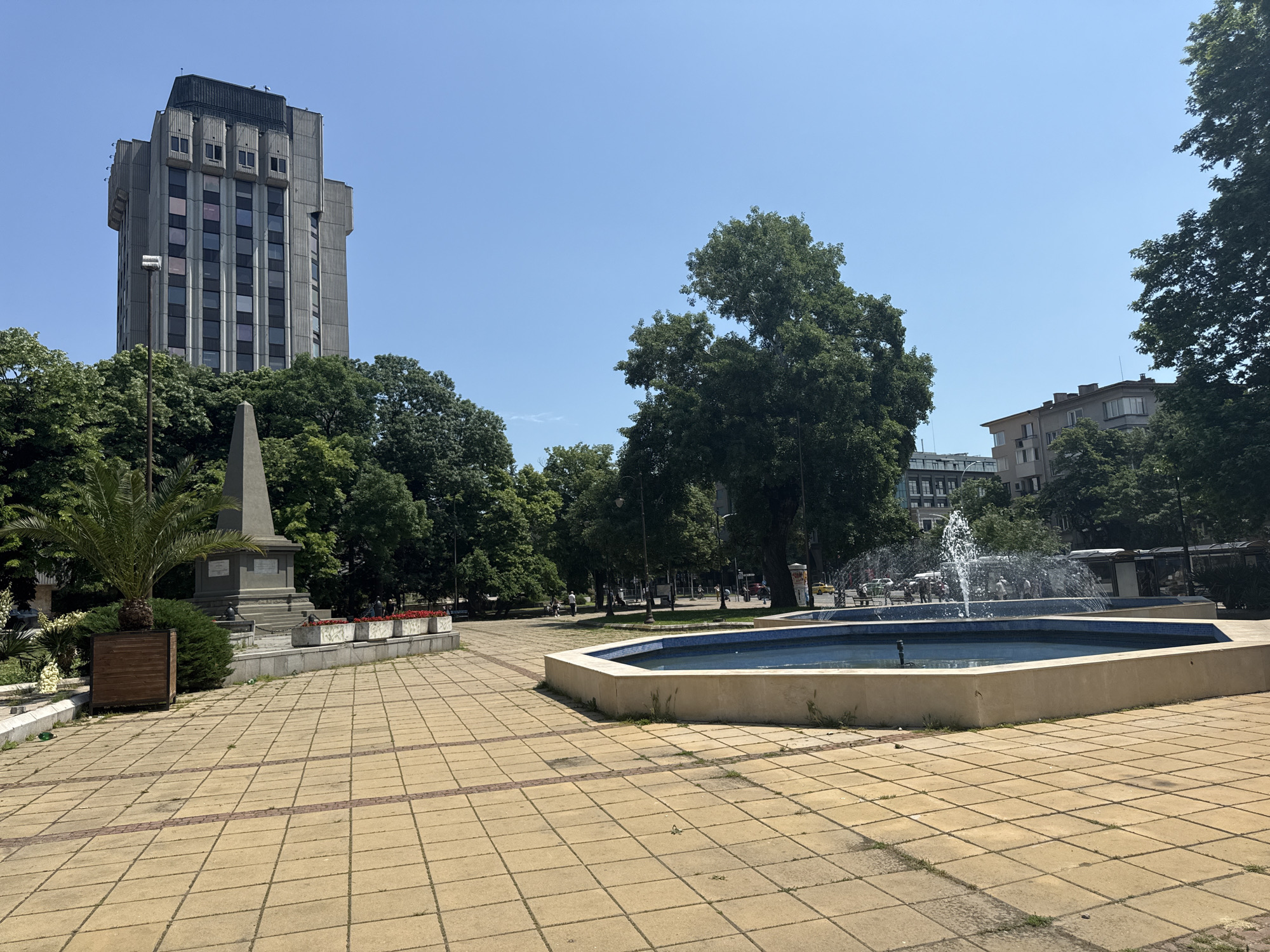 The image captures a sunny urban park or square, paved with light-colored square tiles interspersed with darker, reddish-brown accent tiles forming linear patterns. In the foreground on the right, an octagonal fountain with blue mosaic tiles is actively spraying water. To the left, a stone obelisk-like monument stands on a tiered base, partially framed by a leafy palm tree in a wooden planter box and lush green foliage.
The park is abundant with mature green trees, which fill much of the mid-ground and background. Above the trees on the left, a tall, imposing grey concrete building with a Brutalist architectural style dominates the skyline. It features multiple vertical window rows and distinct, blocky elements near its top. On the far right, beyond the fountain and trees, another residential-style building is partially visible.
In the distant background, near a street hinted at by parked cars and what appears to be a bus stop shelter, a few indistinct figures of people can be seen, suggesting light pedestrian activity. The sky is clear and bright blue, indicating a sunny day, likely mid-morning or early afternoon, given the relatively short shadows. The overall impression is one of a public space in a city, combining natural elements with urban architecture. No discernible text is visible.