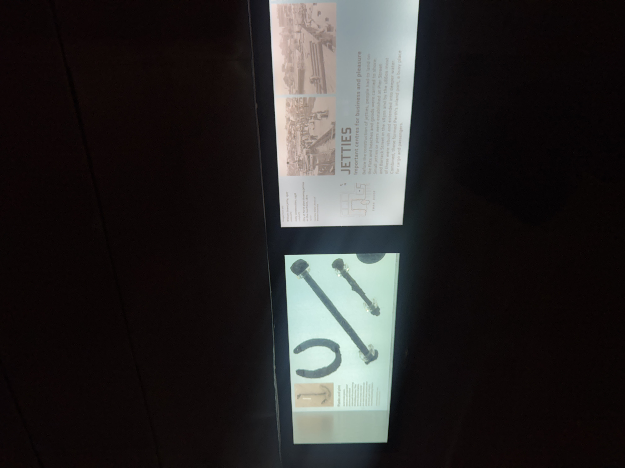 The image captures two vertically oriented, illuminated display panels in what appears to be an exhibition or museum setting, surrounded by darkness. The panels are part of an exhibit detailing the history of jetties.
The upper panel, titled "JETTIES Important centres for business and pleasure," features two historical, sepia-toned photographs depicting bustling jetties, likely on the Swan River, with people and boats. The accompanying text explains that before jetties, people landed on flats and beaches. Small jetties were established at Pier Street and Barrack Street in the 1830s, and by the 1860s, these were rebuilt and extended into deeper water, forming "Perth's inland port, a busy place for cargo and passengers." Other visible text snippets on this panel include "Jetty," "Street jetty," "City of Perth plan showing jetties," and "SWAN RIVER."
The lower panel displays physical artifacts, including three large, weathered metal objects: two long, thick bolts or pins with rounded heads, and a U-shaped object resembling a horseshoe or shackle. A smaller, faded image of an anchor and partial text reading "JIG BOLTS and pins" are also visible at the bottom of this panel. The lighting suggests an indoor, artificial setting, with the exhibits brightly lit against a dark background. The panels are slightly tilted, giving a dynamic perspective.