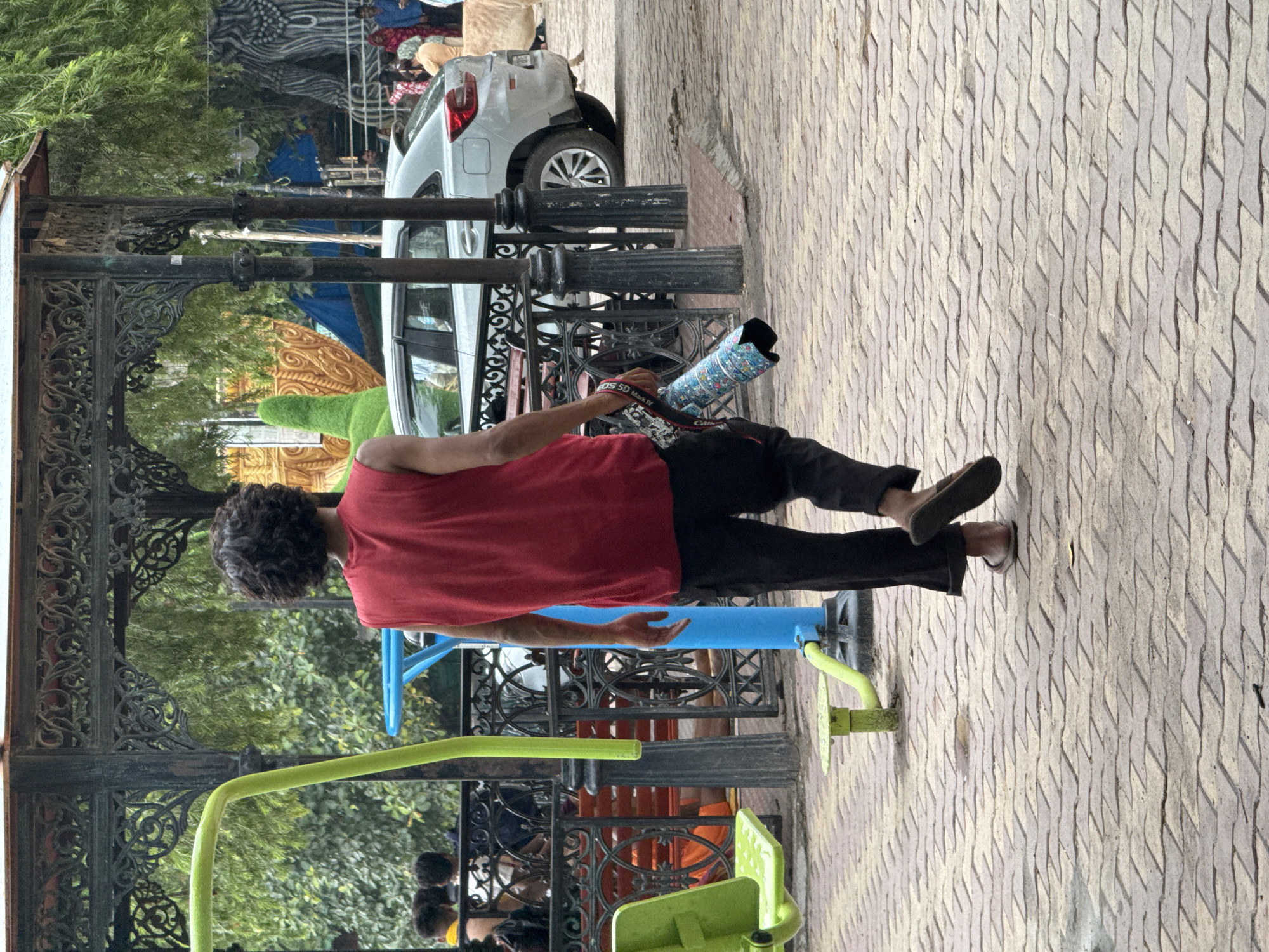 A full-shot, eye-level, outdoor photograph captures a person walking away from the camera on a brick-patterned pathway in Rishikesh, India. The individual, seen from the back, wears a sleeveless red t-shirt and black pants. Their curly, dark hair is visible, and they are carrying a rolled-up item in their right hand, which appears to be a camera bag or a mat, adorned with a colorful, patterned fabric. The person is also wearing dark-colored flip-flops.
The environment suggests a public park or recreational area. In the foreground, to the left, a bright green exercise equipment component is partially visible, along with a blue pole. Behind the person, a silver car is parked on the right side of the pathway. Further back, a decorative metal structure with intricate carvings is evident, suggesting an ornate gazebo or pavilion, with lush green foliage and trees in the background. Other individuals and people are present in the background, indicating a populated area. The lighting suggests it is daytime, with some overcast conditions.