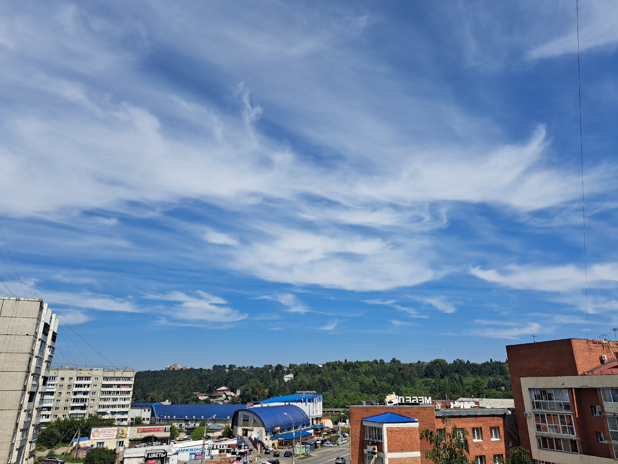 A high-angle, wide shot shows a city skyline under a partly cloudy blue sky. In the foreground, to the left, is a multi-story apartment building with a gray, textured facade. To the right, is a red brick building with a white trim, featuring multiple windows and balconies. In the middle ground, there are several commercial buildings with blue roofs, and a tree-lined hill with houses visible in the distance. Several cars are visible on a road in front of the commercial buildings. The sky is filled with wispy white clouds. The image appears to be taken in Irkutsk, Russia, given the architectural style of the buildings and the visible Cyrillic text on the signs.