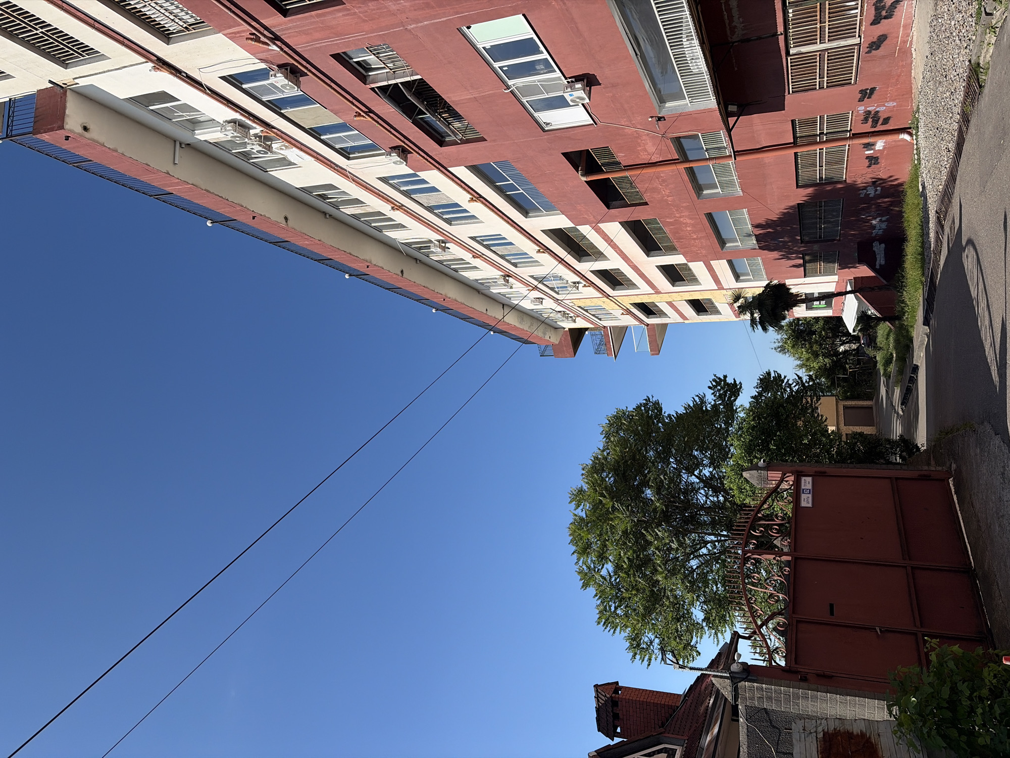 This image, captured in Sochi, Russia, depicts an outdoor scene under a clear blue sky with bright daylight. A multi-story building with alternating red and white facades dominates the right side of the frame, angled upwards. Its red sections display signs of weathering and minor graffiti, while the white sections feature numerous rectangular windows, some equipped with external air conditioning units. Black metal railings are present on upper-floor balconies. To the left, a large, dark red metal gate, adorned with decorative scrollwork, stands mostly closed. A blue sign reading "ТОБОЛУРГ СПОРТ ЦЕНТР" (Tobolurg Sport Center) is affixed to the gate. Adjacent to the gate on the far left, a portion of a house with a red-tiled roof and a brick chimney is visible. A prominent green-leafed deciduous tree grows behind the gate, and a slender palm tree is seen against the lower-left facade of the large building. Two thin, dark utility lines traverse the upper sky diagonally. An asphalt road, exhibiting minor surface wear and a storm drain, occupies the foreground. Strong shadows from buildings and trees are cast across the road and the lower building facade. No individuals are present.