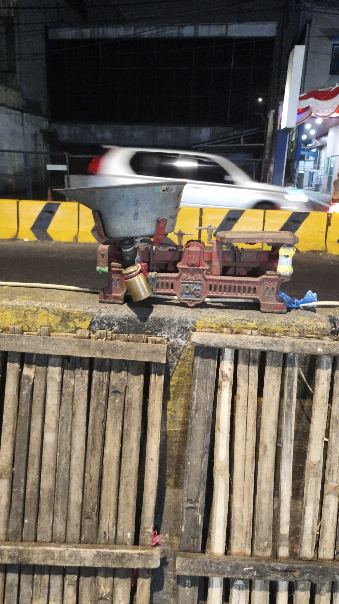 In South Jakarta, Indonesia, a vintage red scale with a gray basin sits on a concrete barrier. A cylindrical brass weight is attached to the side of the scale. Behind the scale, a white car is blurred due to motion, suggesting it is driving past. The barrier is marked with yellow and black diagonal stripes. In the foreground, a fence constructed from vertical wooden planks is visible. The scene is set at night, with artificial lighting illuminating the area.
