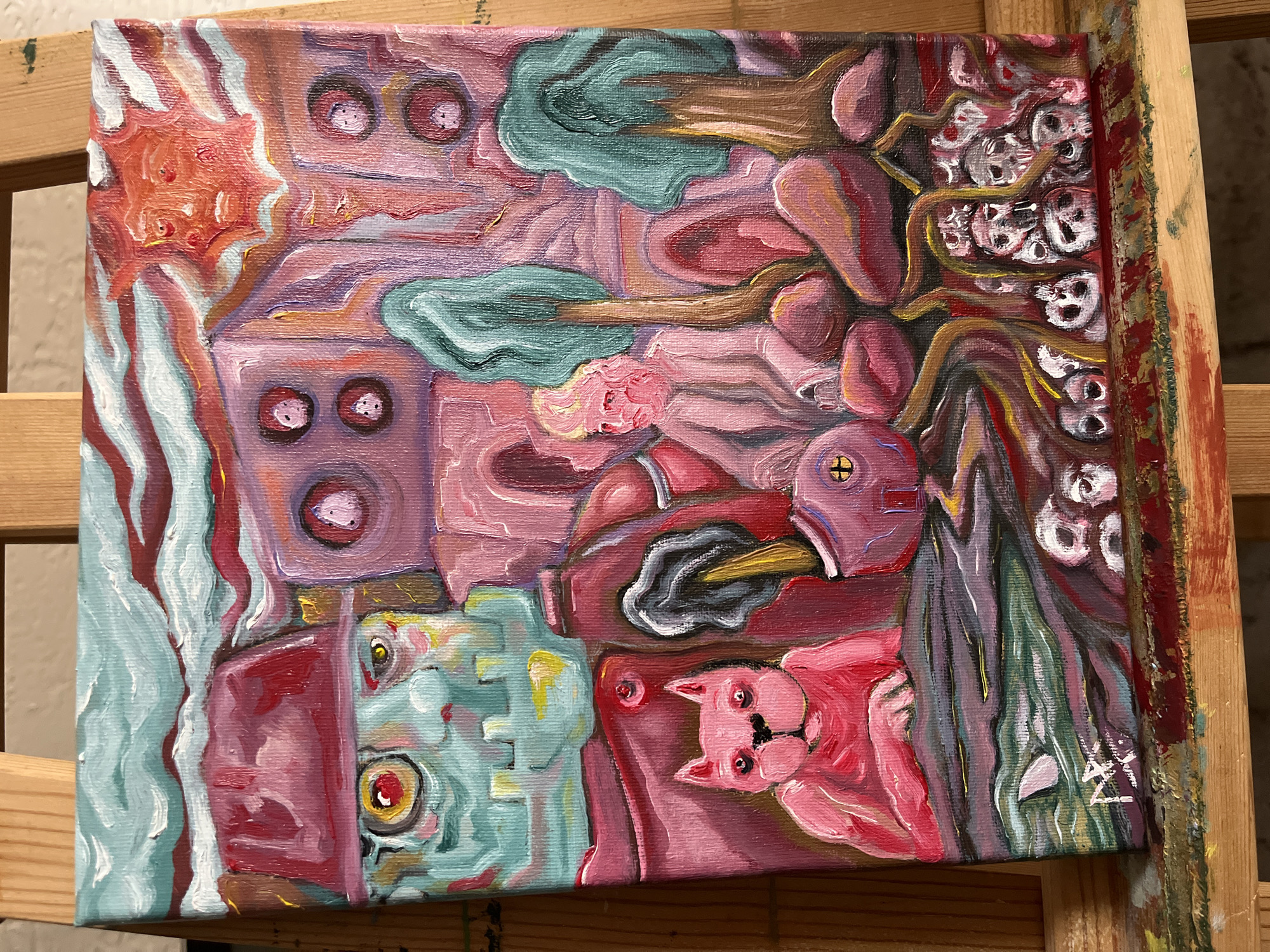 The image is an oil painting on canvas. The scene depicts an abstract, surreal environment with various elements. A blue and red structure is on the left, with a greenish-blue creature with a prominent yellow eye next to it. A pink cat-like figure with red eyes is in the lower right corner. Above the cat is a red object with a black section and a golden beak-like extension. In the upper right, a cluster of small skulls is intertwined with brown root-like structures. The upper area features abstract shapes in shades of pink, lavender, and teal. The painting is mounted on a wooden easel.